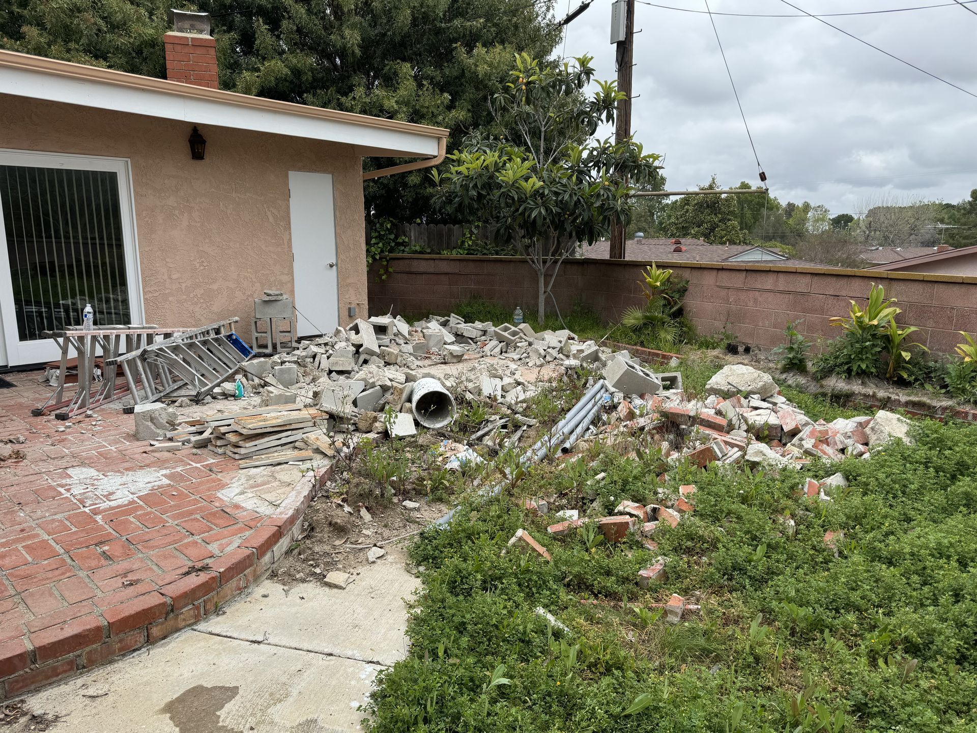 Backyard with piles of construction debris, a broken concrete walkway, and a house patio at left