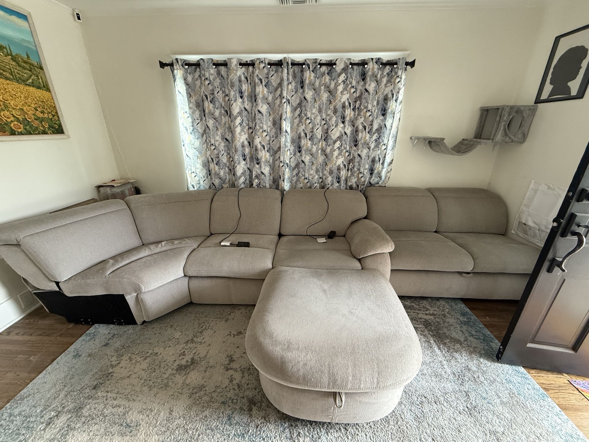 Beige sectional sofa with ottoman in a living room, centered under a window with patterned curtains.