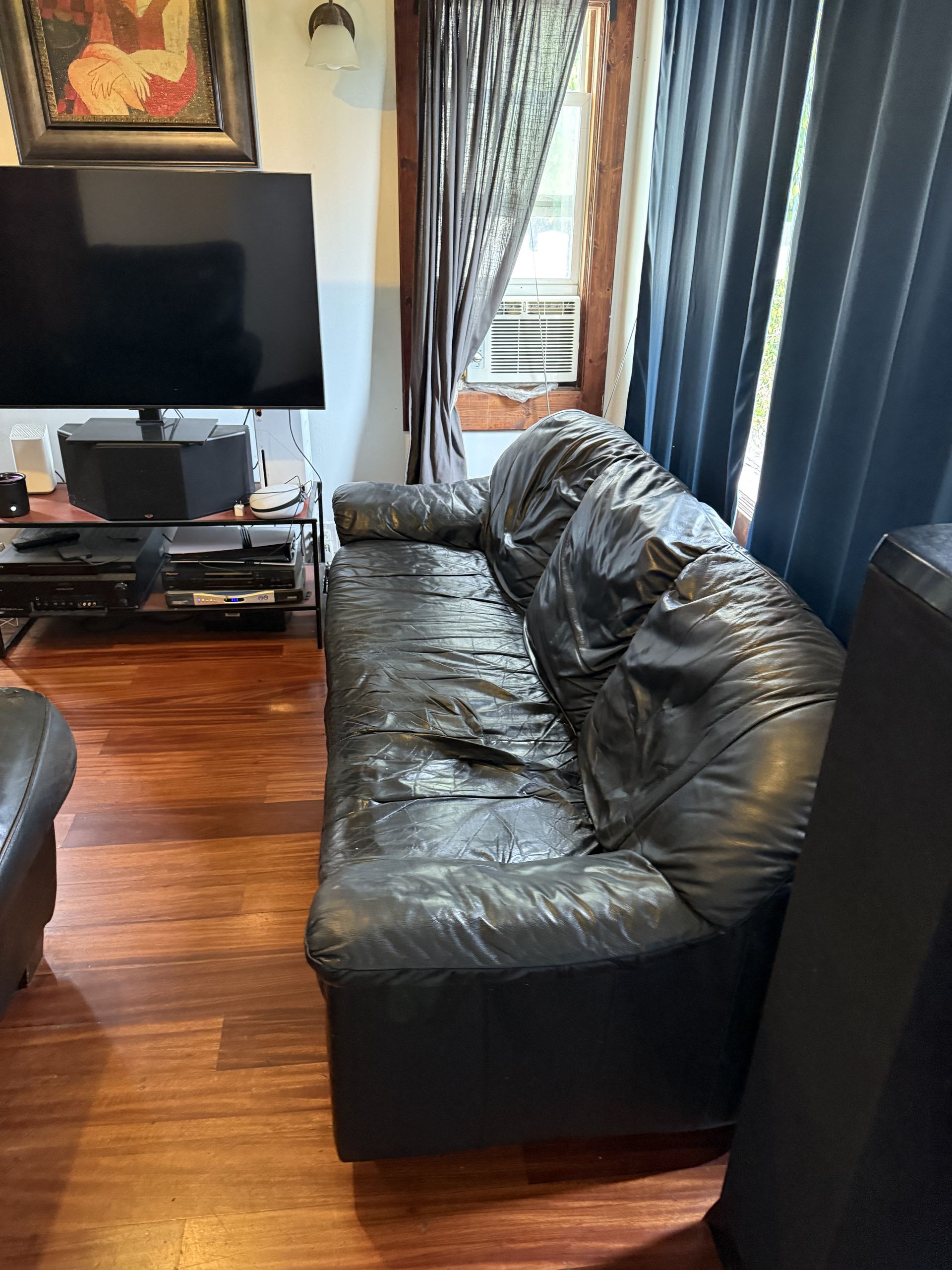 Living room with a black leather couch, TV, hardwood floor, and dark curtains.