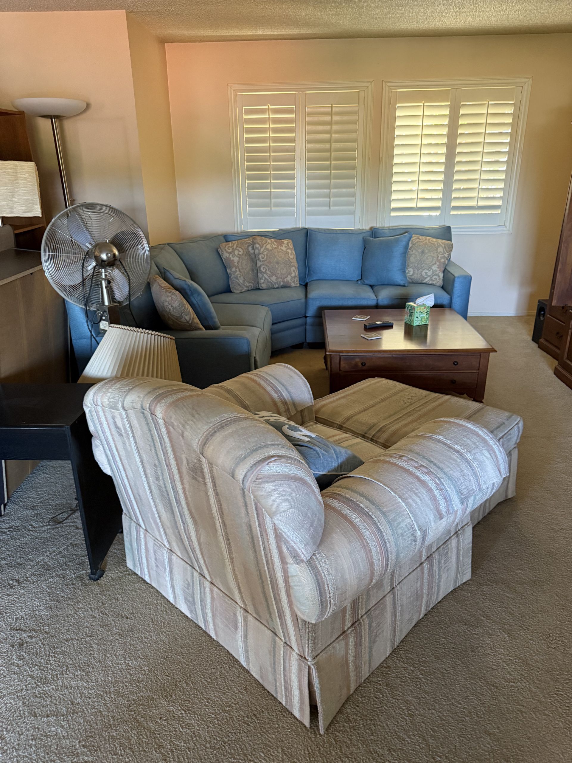 Cozy living room with beige sofa, patterned armchair, coffee table, fan, and white window shutters