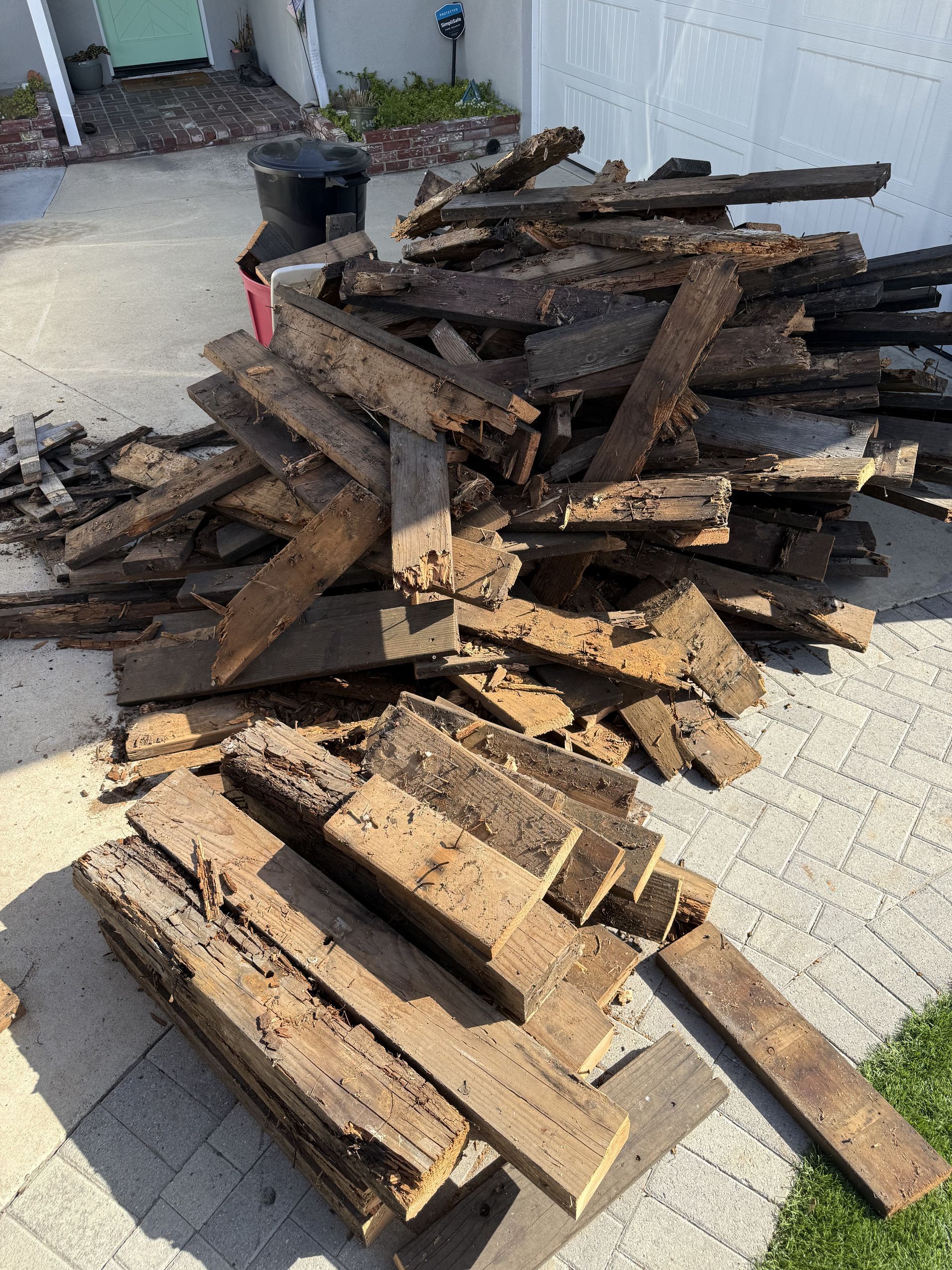 Pile of rough-cut lumber stacked on a paved driveway near a house