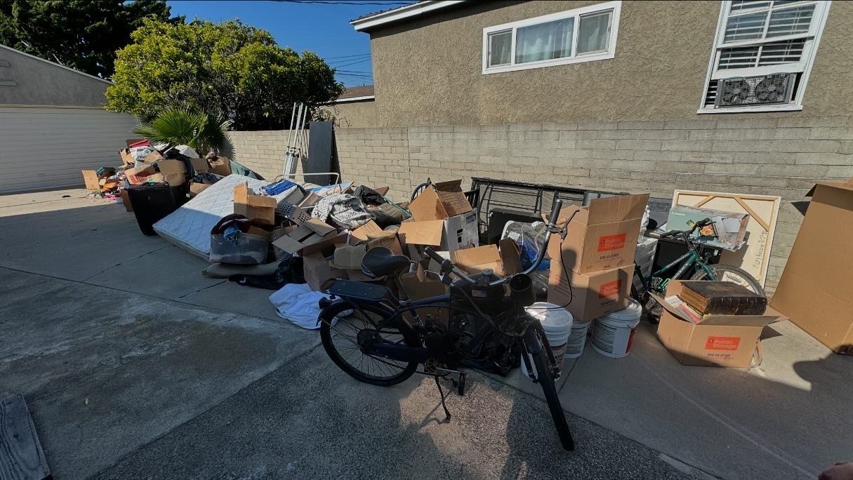 Bicycle and assorted boxes piled on a driveway beside a house, suggesting junk or moving day cleanup.