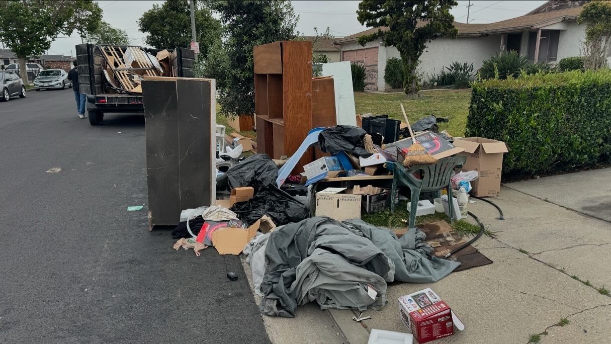 Piled curbside trash and discarded furniture on a residential street near houses and hedges