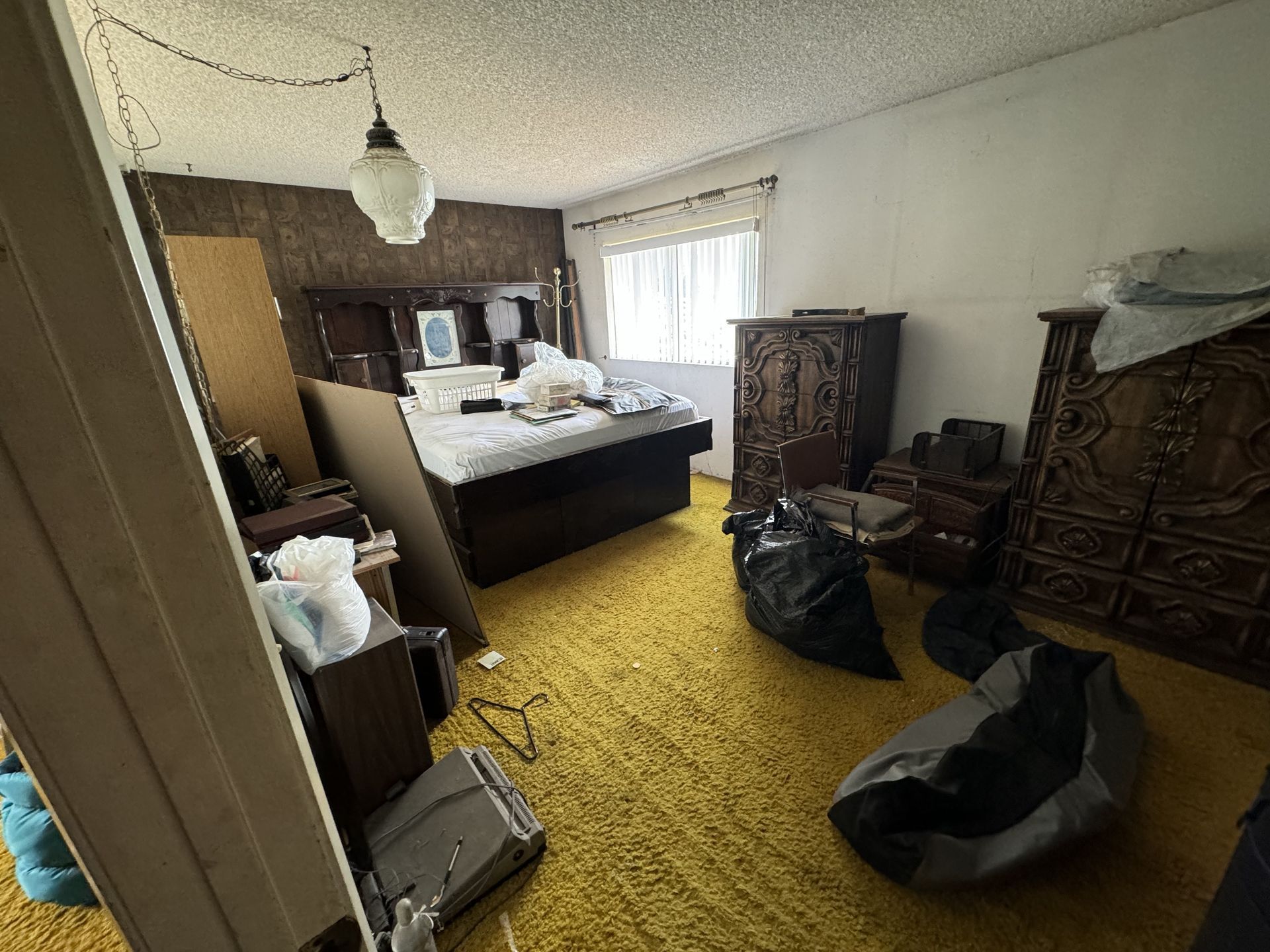 Cluttered bedroom with a bed, cabinets, bags on the floor, and a window with sheer curtains.