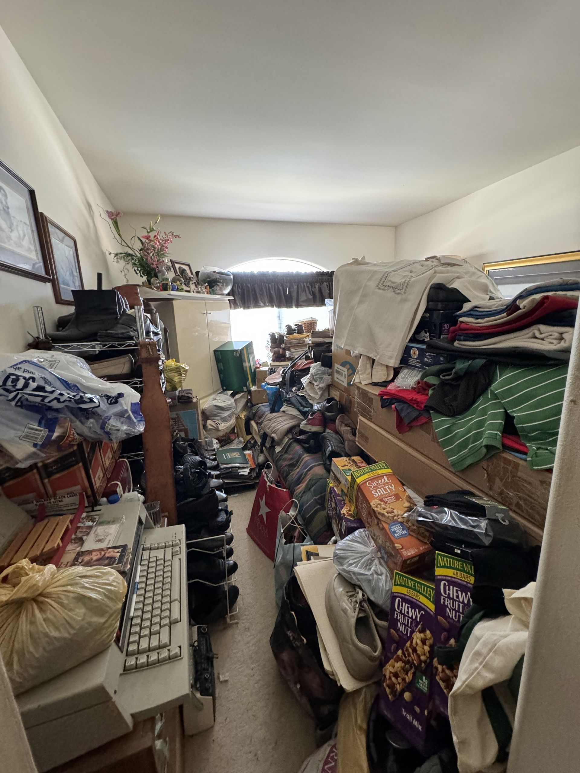 Cluttered room packed with overflowing clothes, boxes, and household items on shelves and the floor.