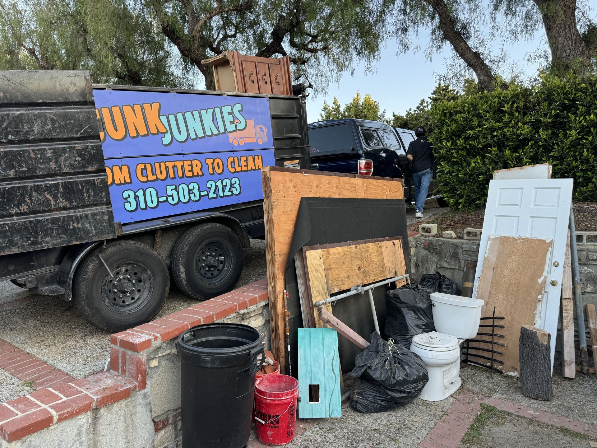 Junk removal trailer with cluttered debris, signs, bins, and a white door outdoors by a brick wall