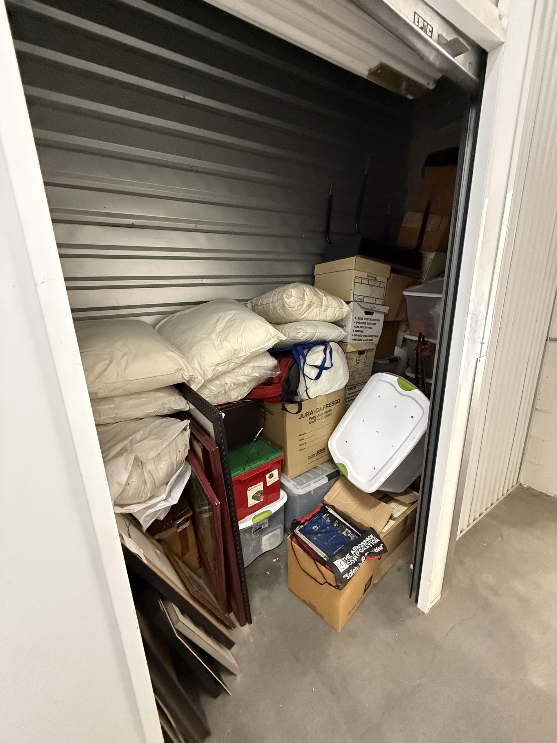 Storage closet with shelves, boxes, bags, and rolled mats piled in a narrow space on a gray floor.