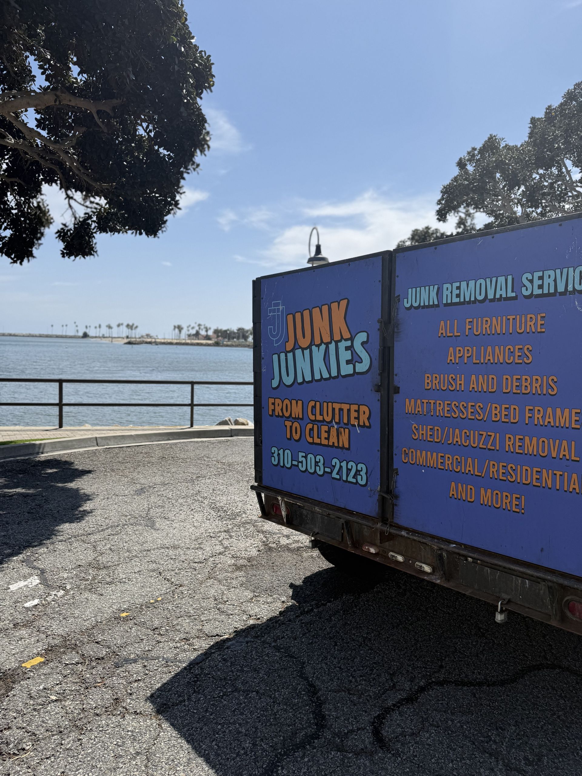 Blue JUNK JUNKIES trailer by a waterfront, with white service text and trees on a gravel lot.