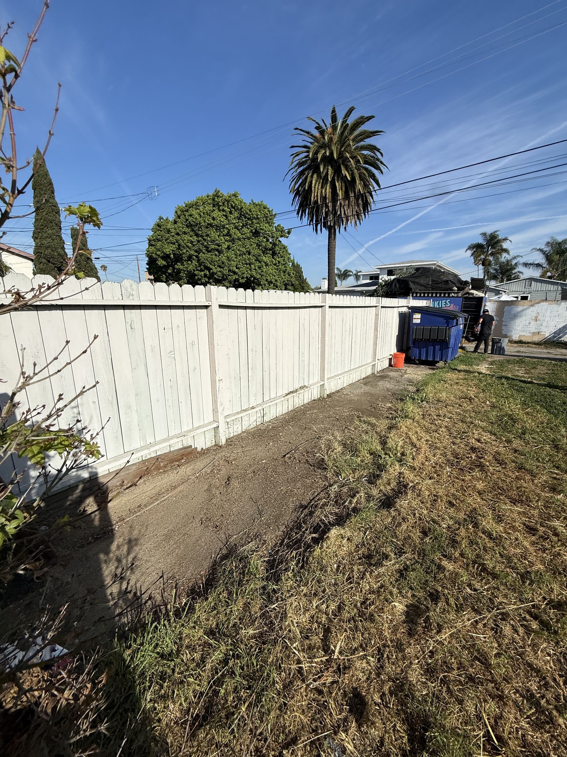 Narrow dirt path beside a white fence, with a palm tree and blue dumpster under a clear blue sky.