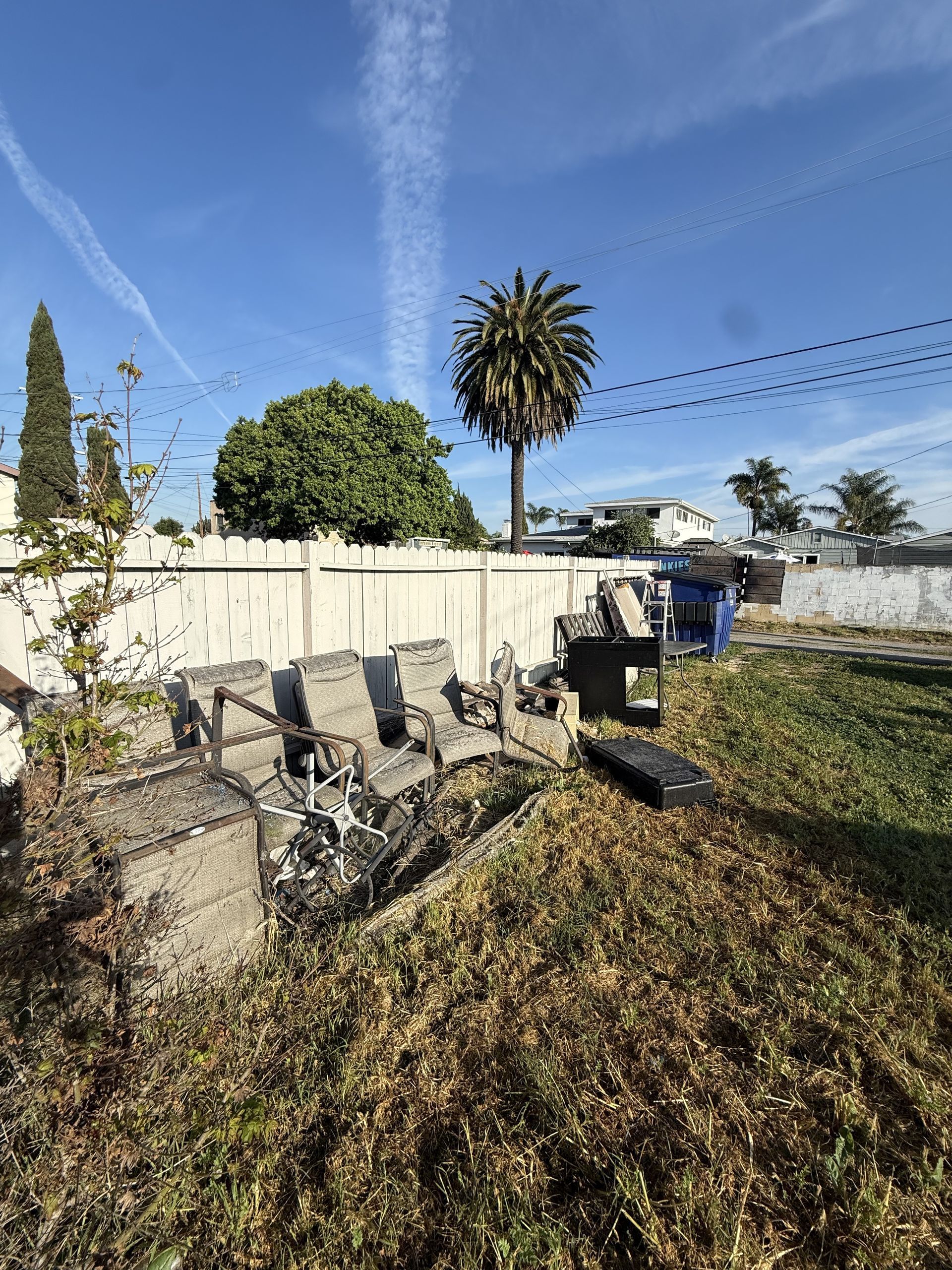 Backyard with charred debris, a white fence, and a palm tree under a clear blue sky