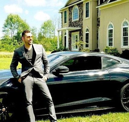 A man in a suit is standing next to a black sports car in front of a house.