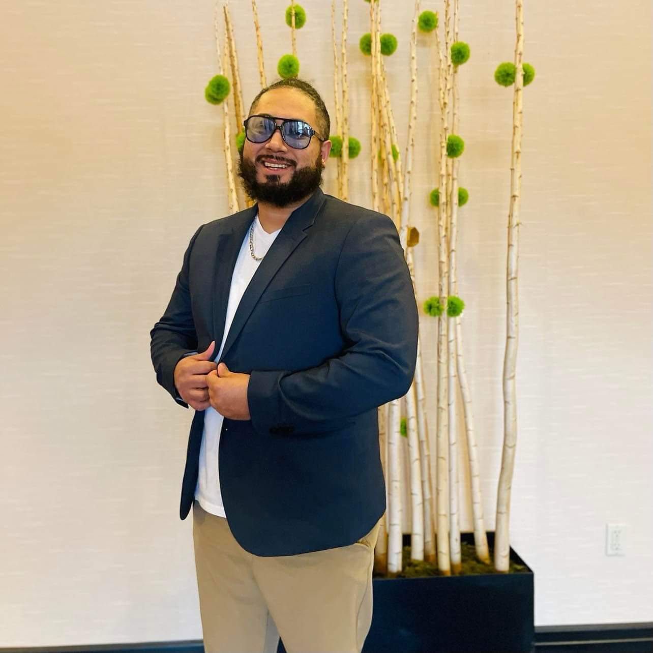 A man in a suit and sunglasses is standing in front of a potted plant.