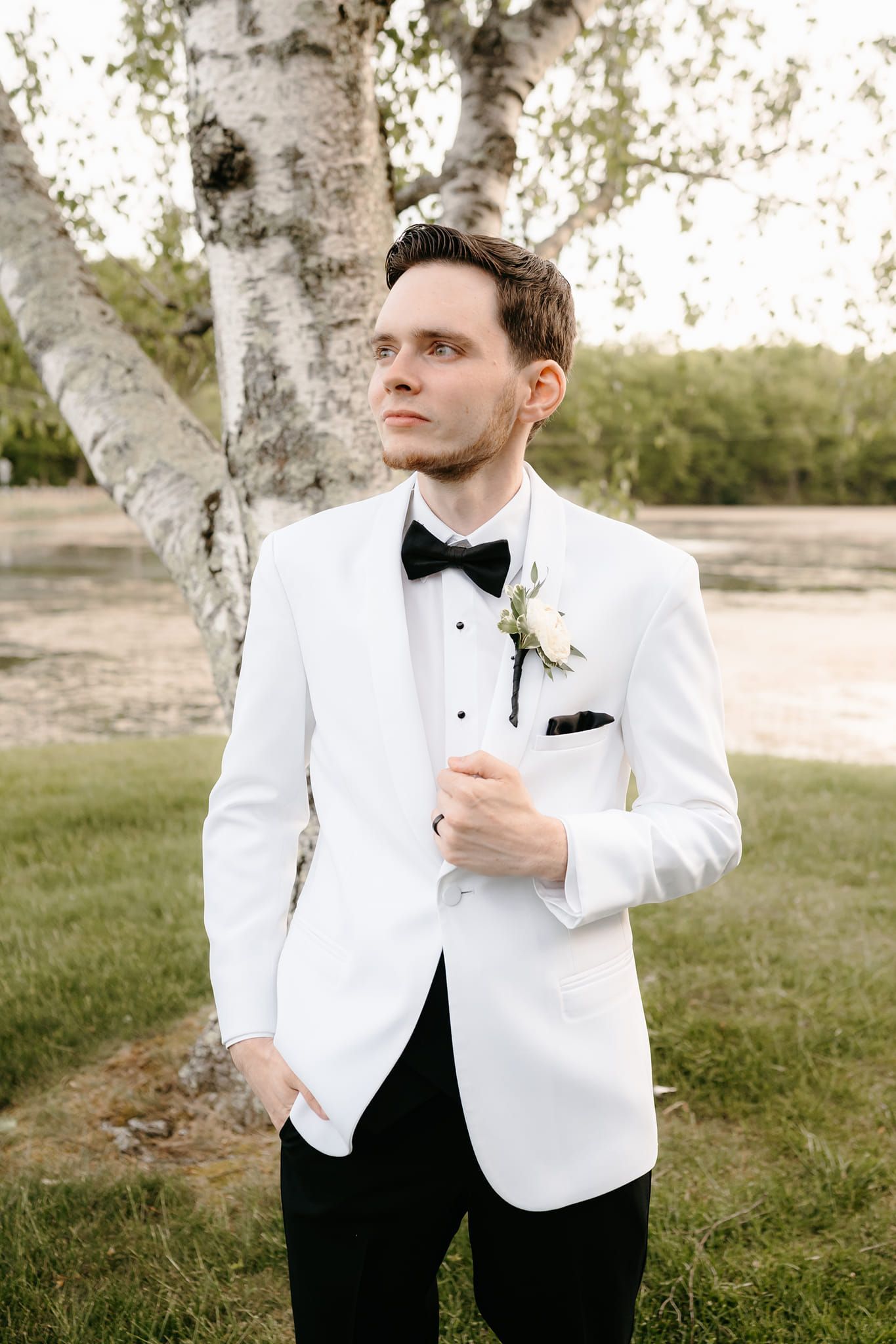 A man in a white tuxedo and bow tie is standing in front of a tree.