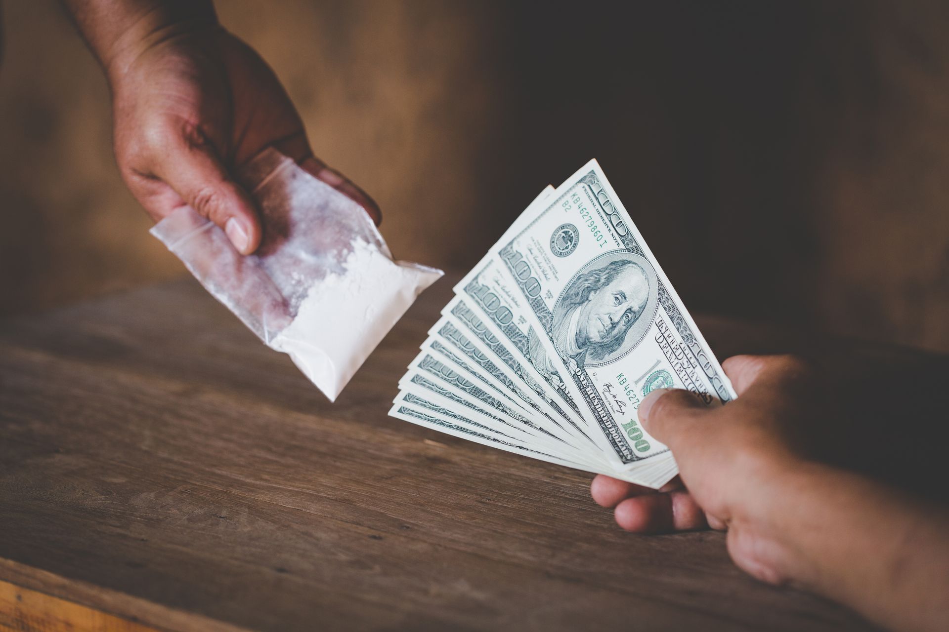 Hands exchanging a bag of white powder for a stack of US dollar bills on a wooden surface.