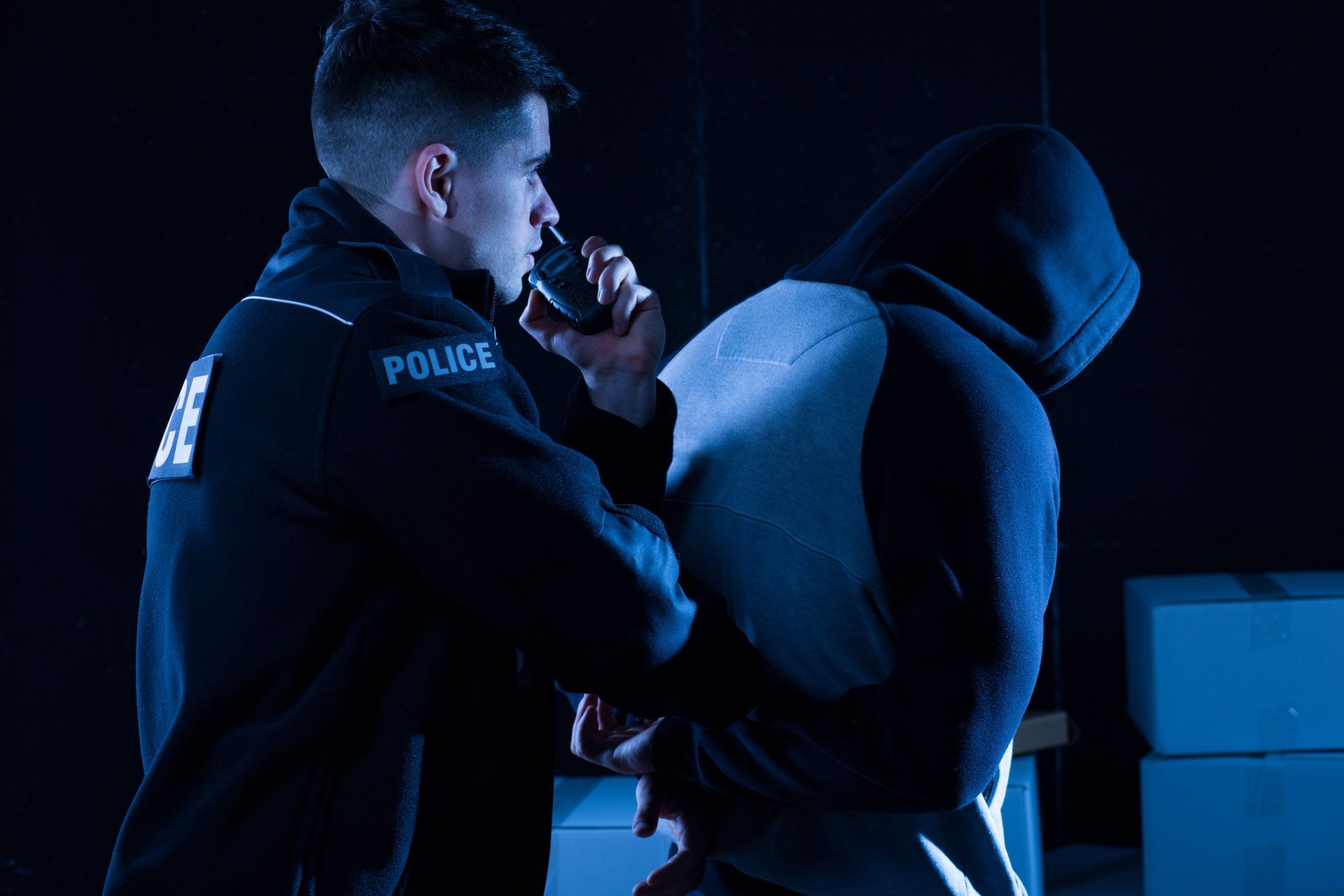 A police officer speaks into a radio while handcuffing a person wearing a hooded sweatshirt in a dimly lit room.
