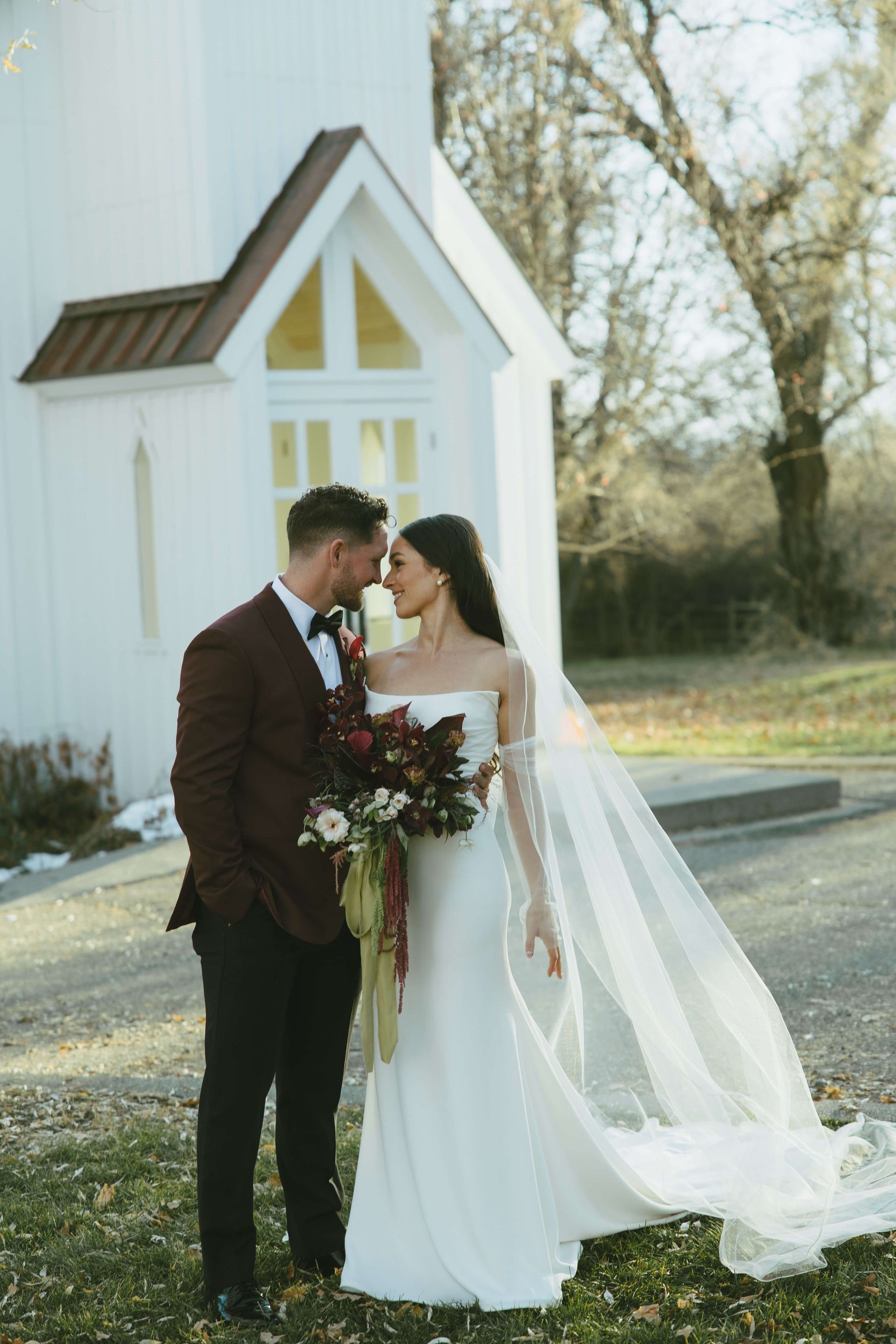 Bride and groom kissing beside a white chapel, bride in flowing veil holding bouquet