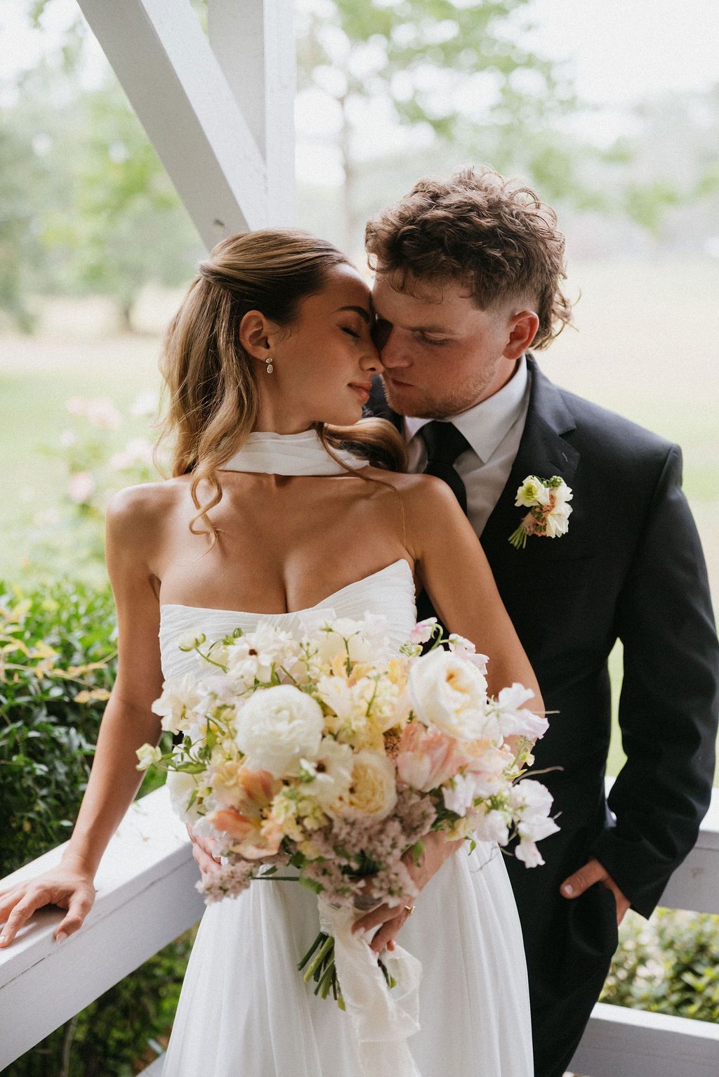 Wedding couple embracing on a balcony, bride holding a bouquet and groom in a black suit kissing her forehead