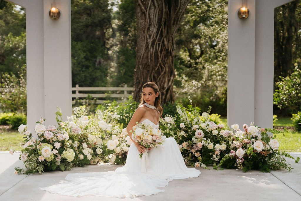 Bride in a white gown seated among white floral arrangements in a garden pavilion.