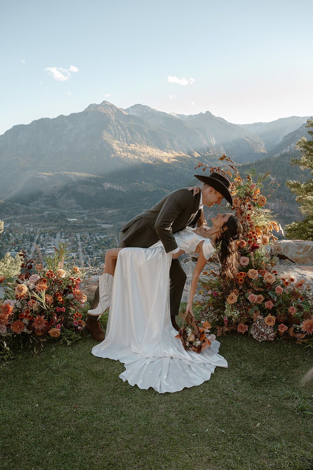 Bride in white dress bending among floral arrangements on a mountain overlook at sunset