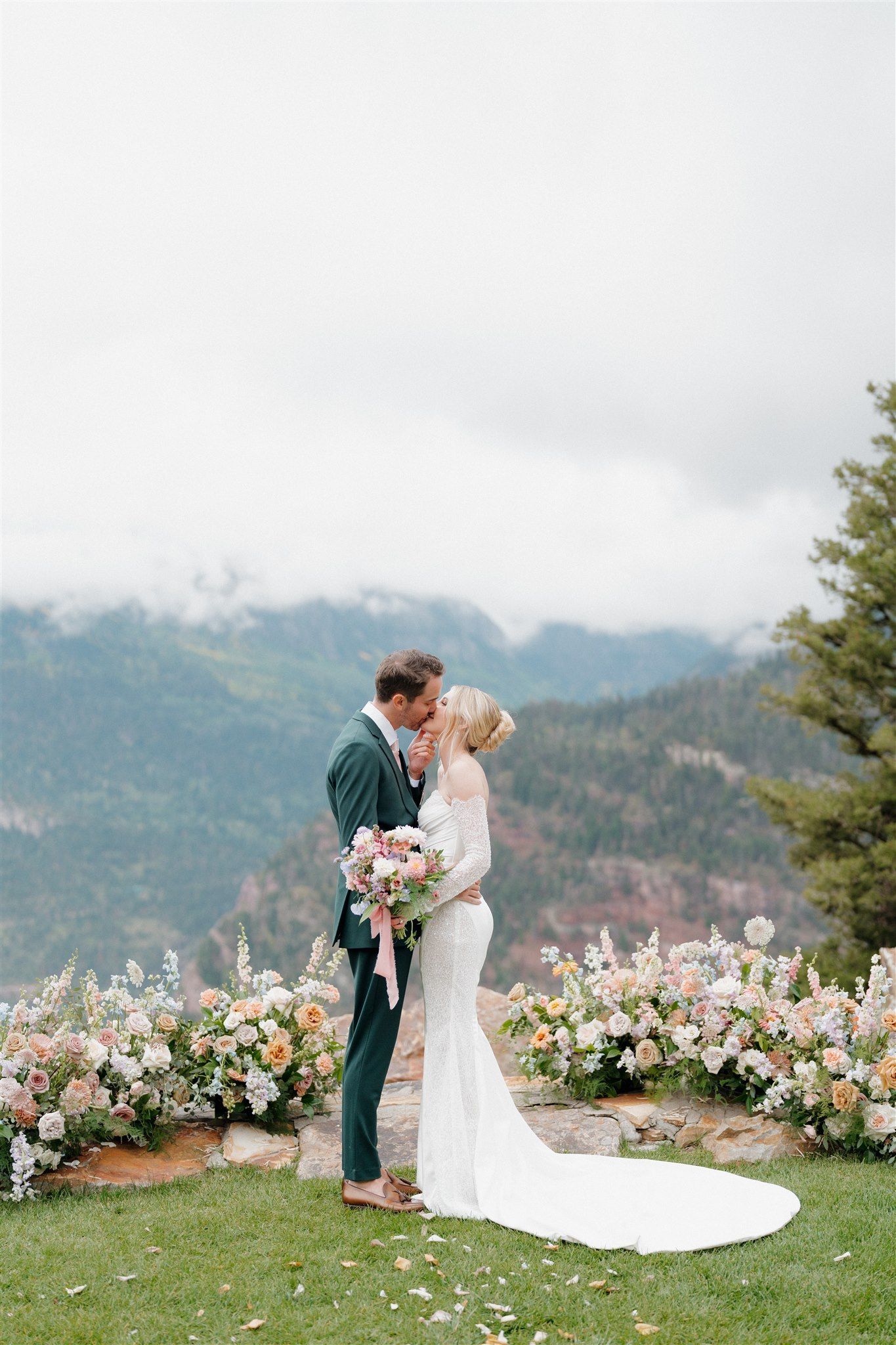 Couple kissing, surrounded by flowers, with mountain view in the background.