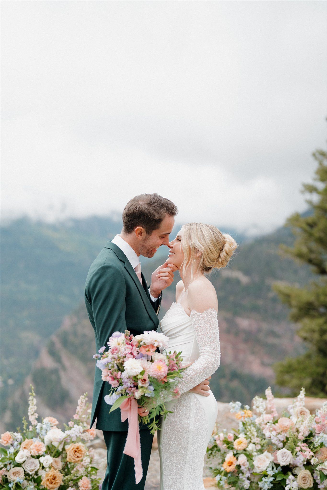 Couple on a mountaintop, embracing. Bride in white dress, groom in green suit. Flowers and scenic background.