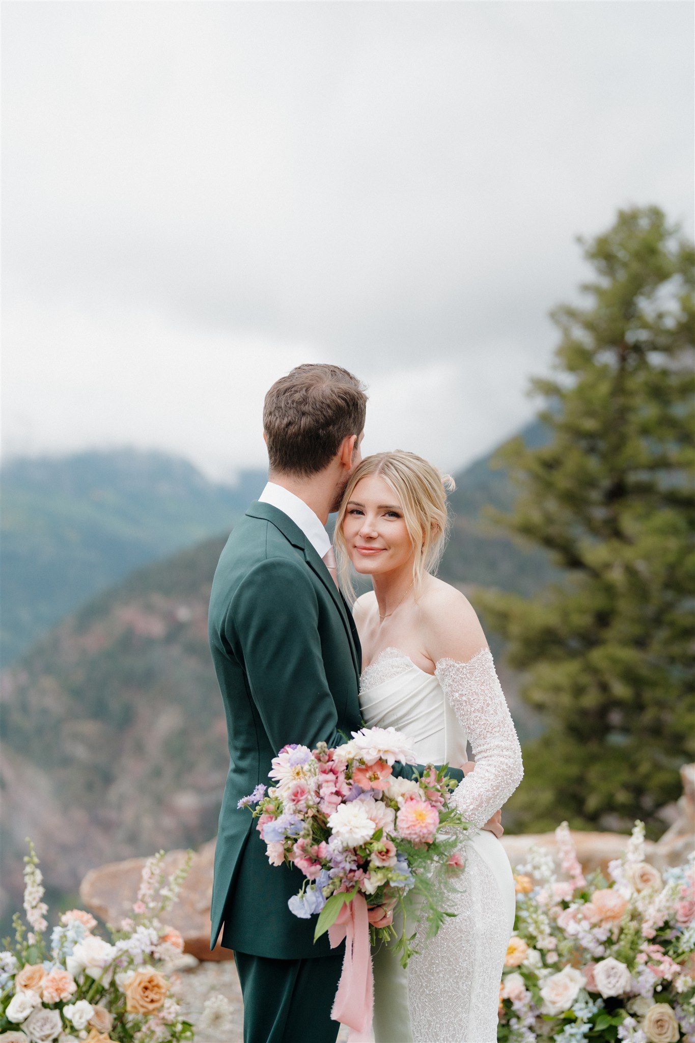 Bride and groom embrace outdoors, holding bouquet. Mountains in background, cloudy sky.