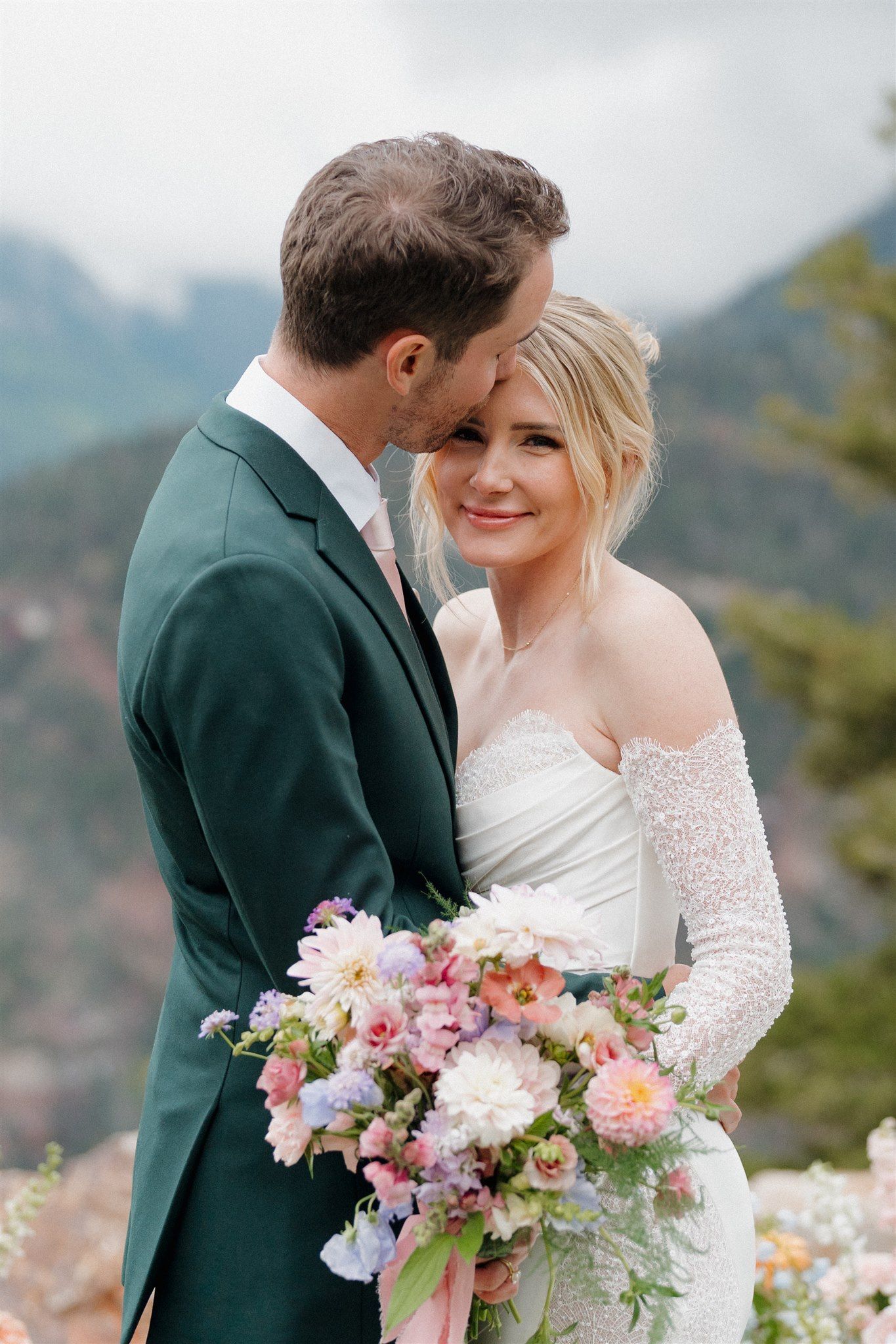 Bride and groom embracing, holding bouquet, outdoors, mountains in background. Bride in white dress, groom in green suit.