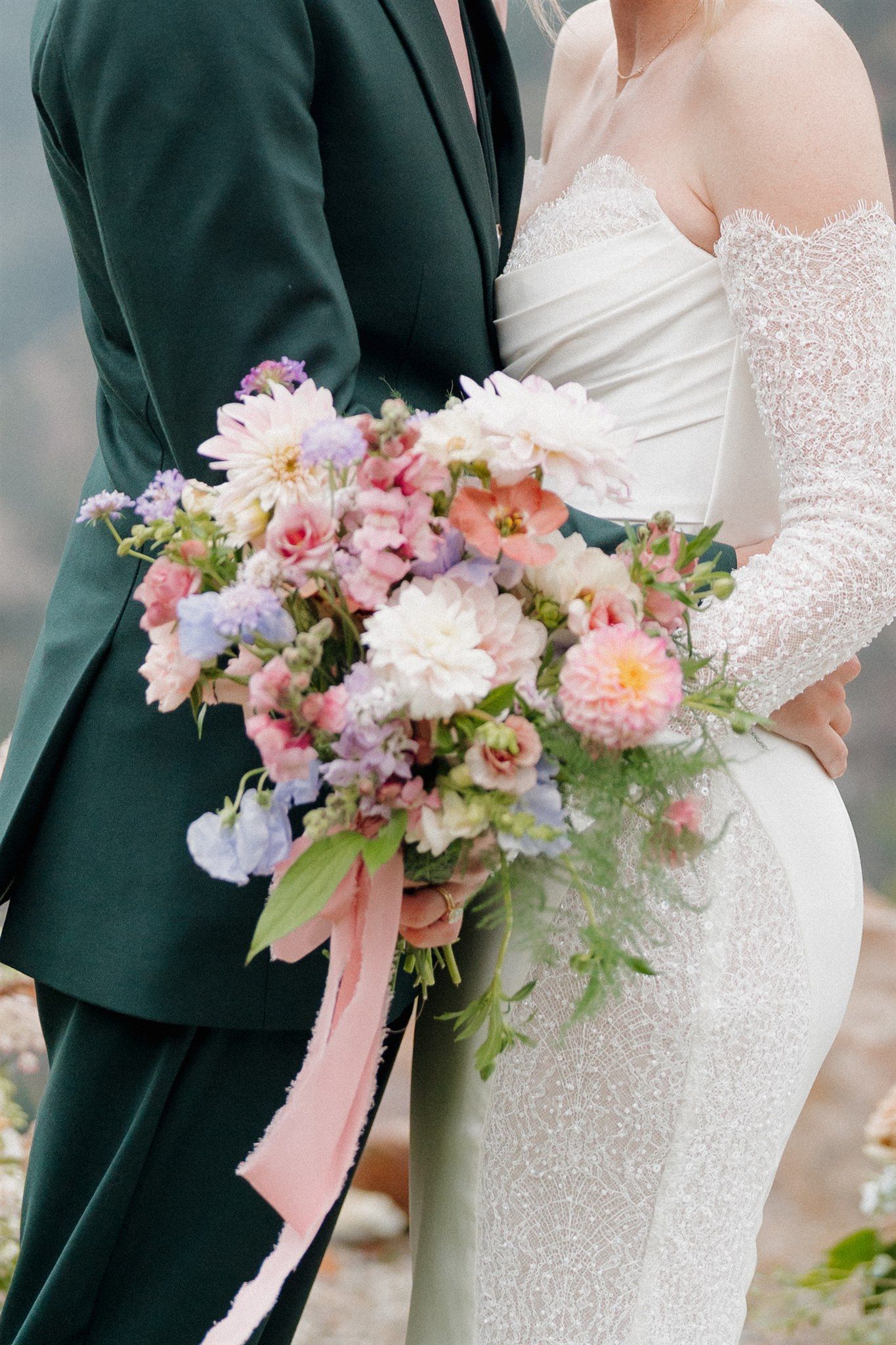 Bride in white gown and groom in green suit holding a pastel flower bouquet, outdoors.