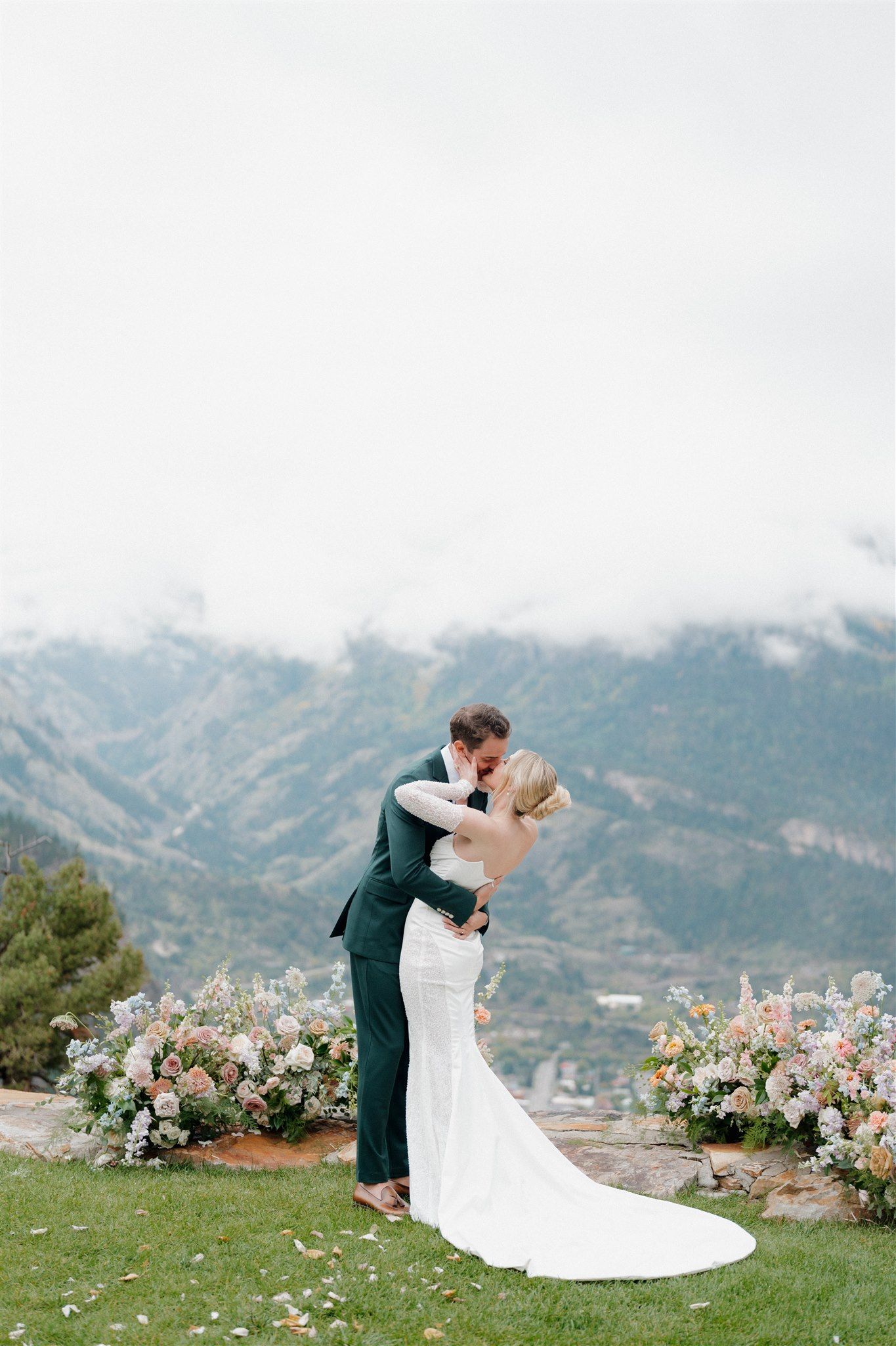 Couple kissing, man in dark suit, woman in white dress, mountain background, flowers on either side.