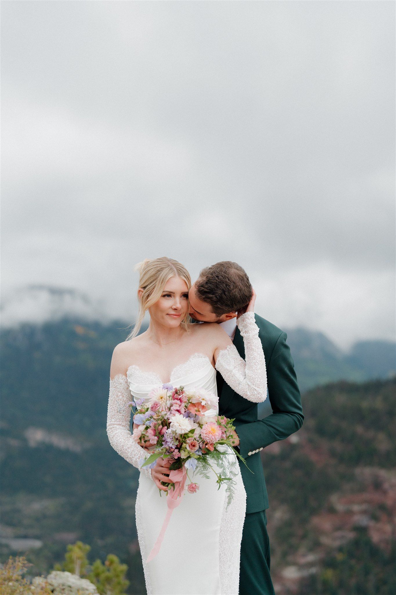 Bride and groom embrace outdoors, groom kisses bride's neck. Mountain landscape with cloudy sky.