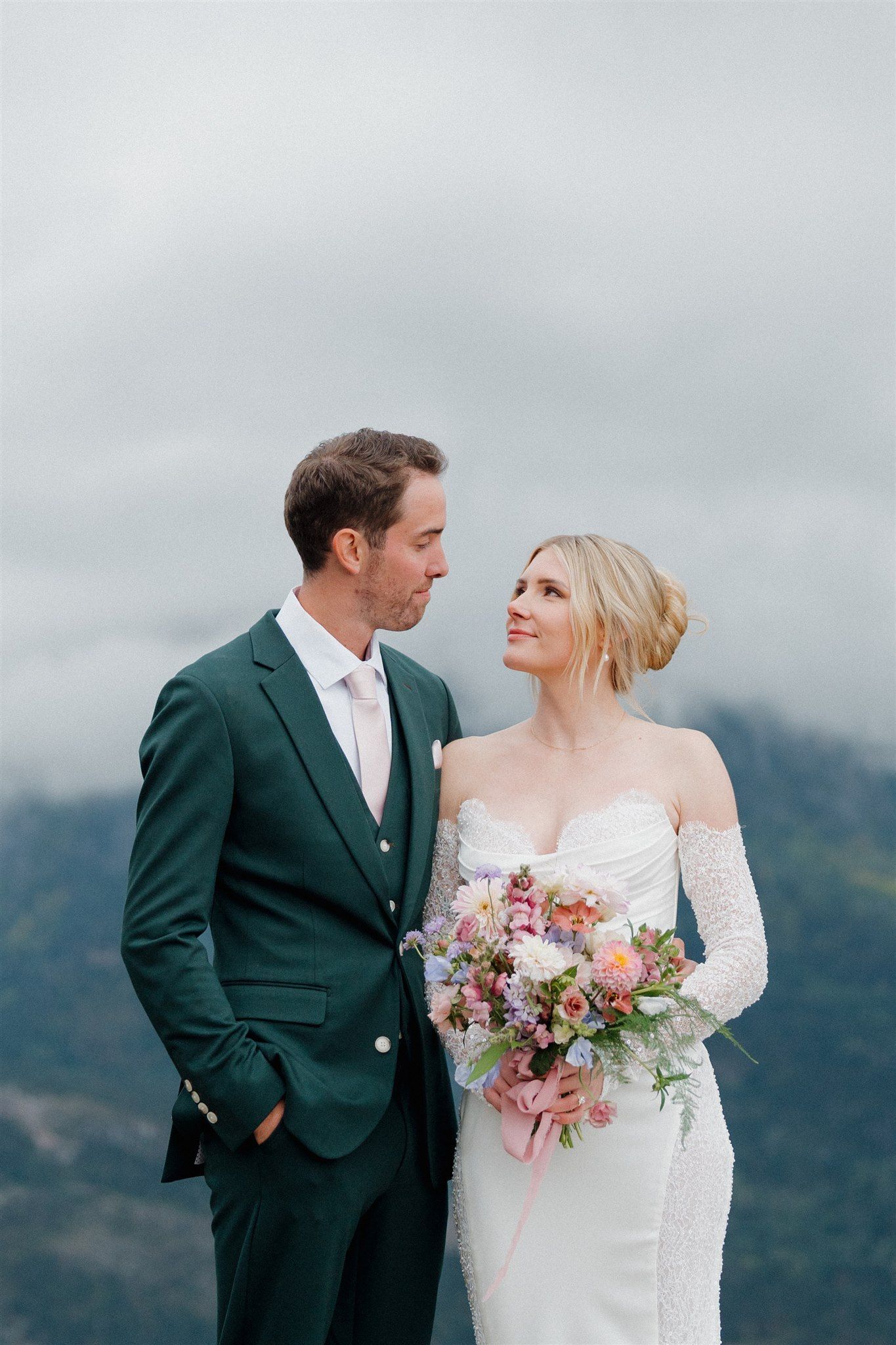 Couple in wedding attire gazing at each other; the man in a green suit, the woman in a white gown holding a bouquet; mountains in background.