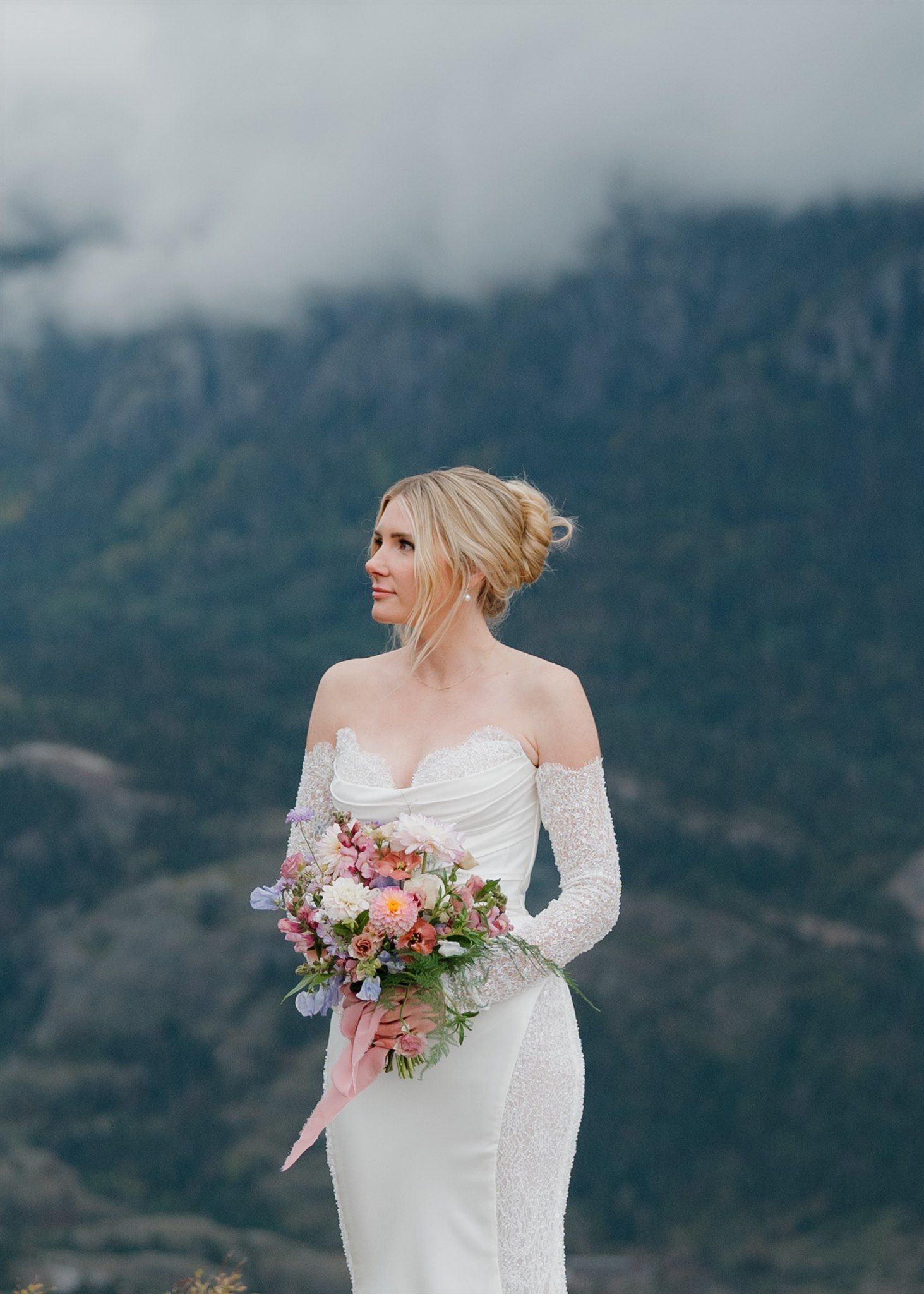 Bride in white dress holding flowers, overlooking a mountain range.