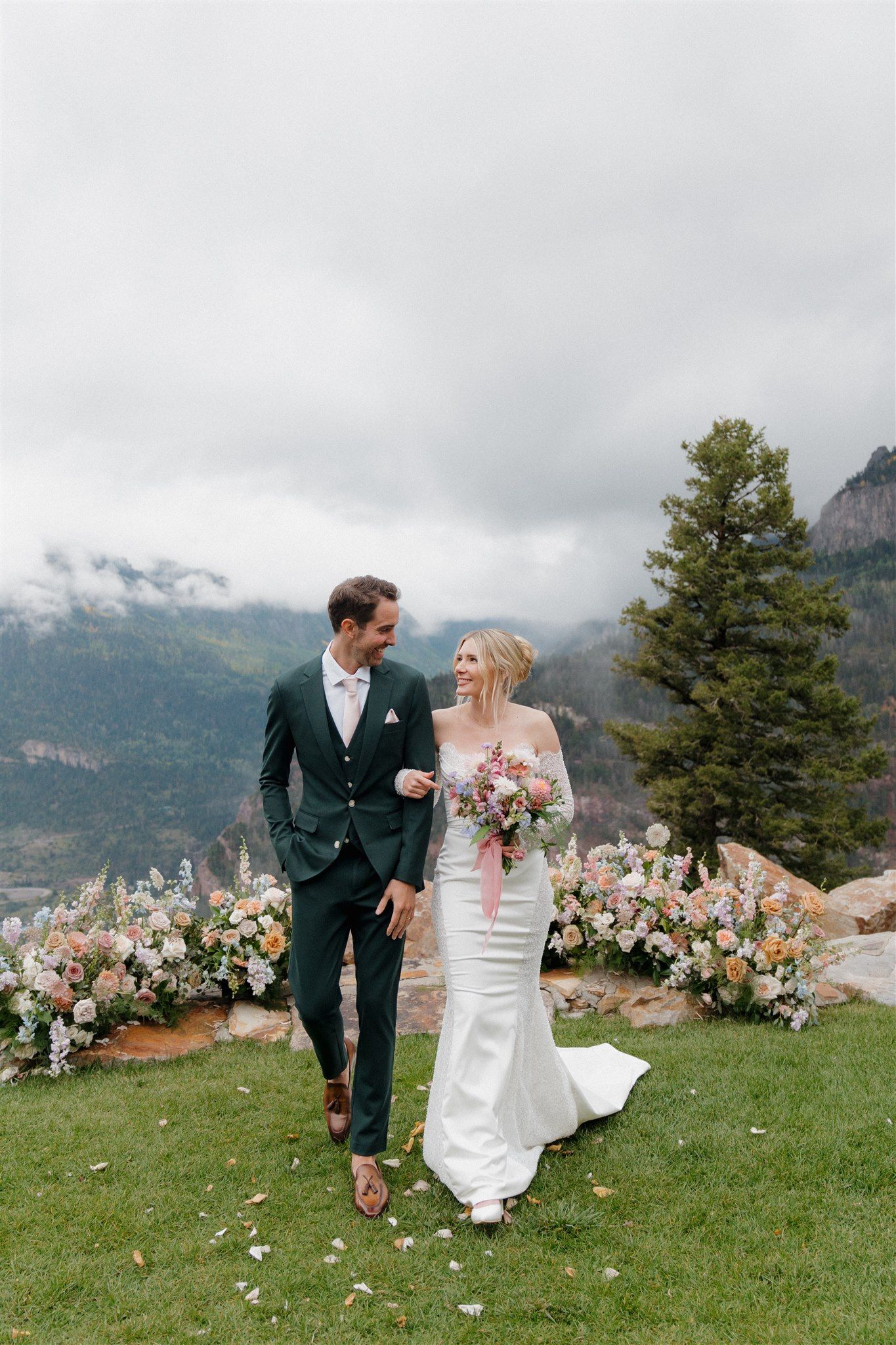 Newly married couple walking arm in arm; scenic mountain backdrop, bride in white dress, groom in green suit.