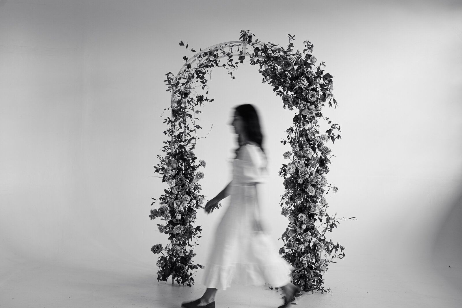 Woman in white dress walks through floral arch in a studio.