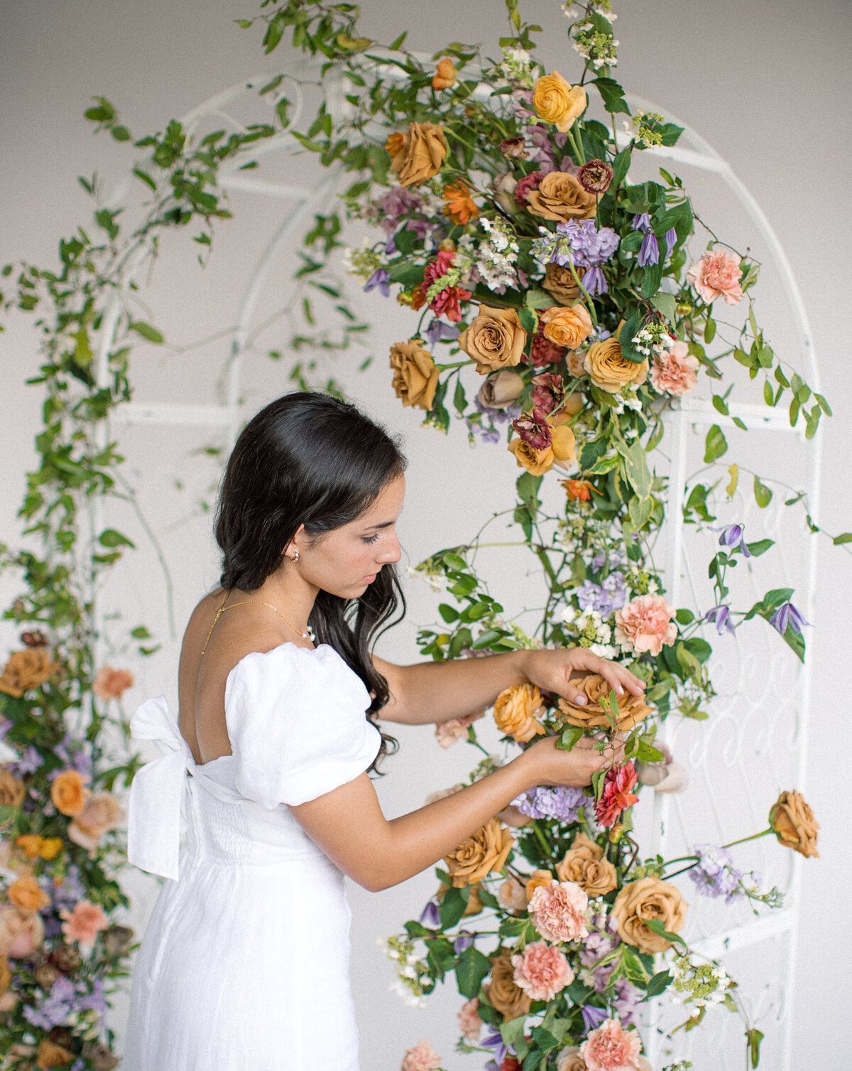 Woman arranging flowers on a white archway covered in greenery and colorful blooms.