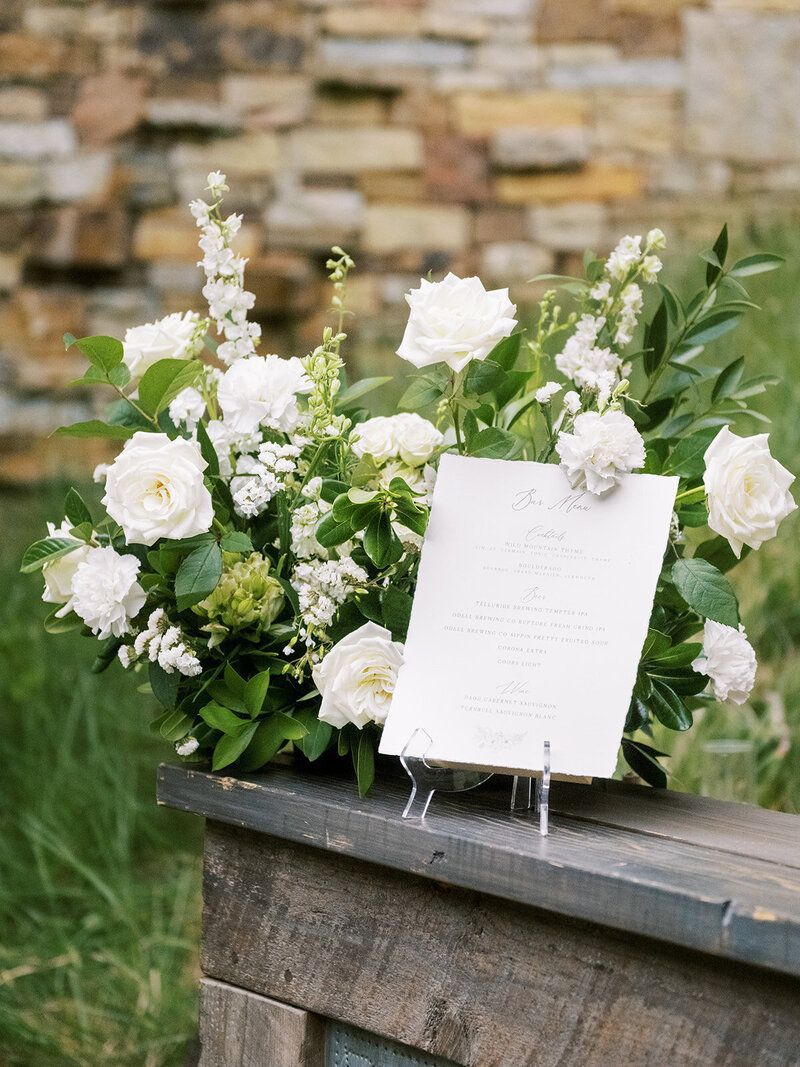 A sign sitting on top of a wooden table next to a bouquet of white flowers.