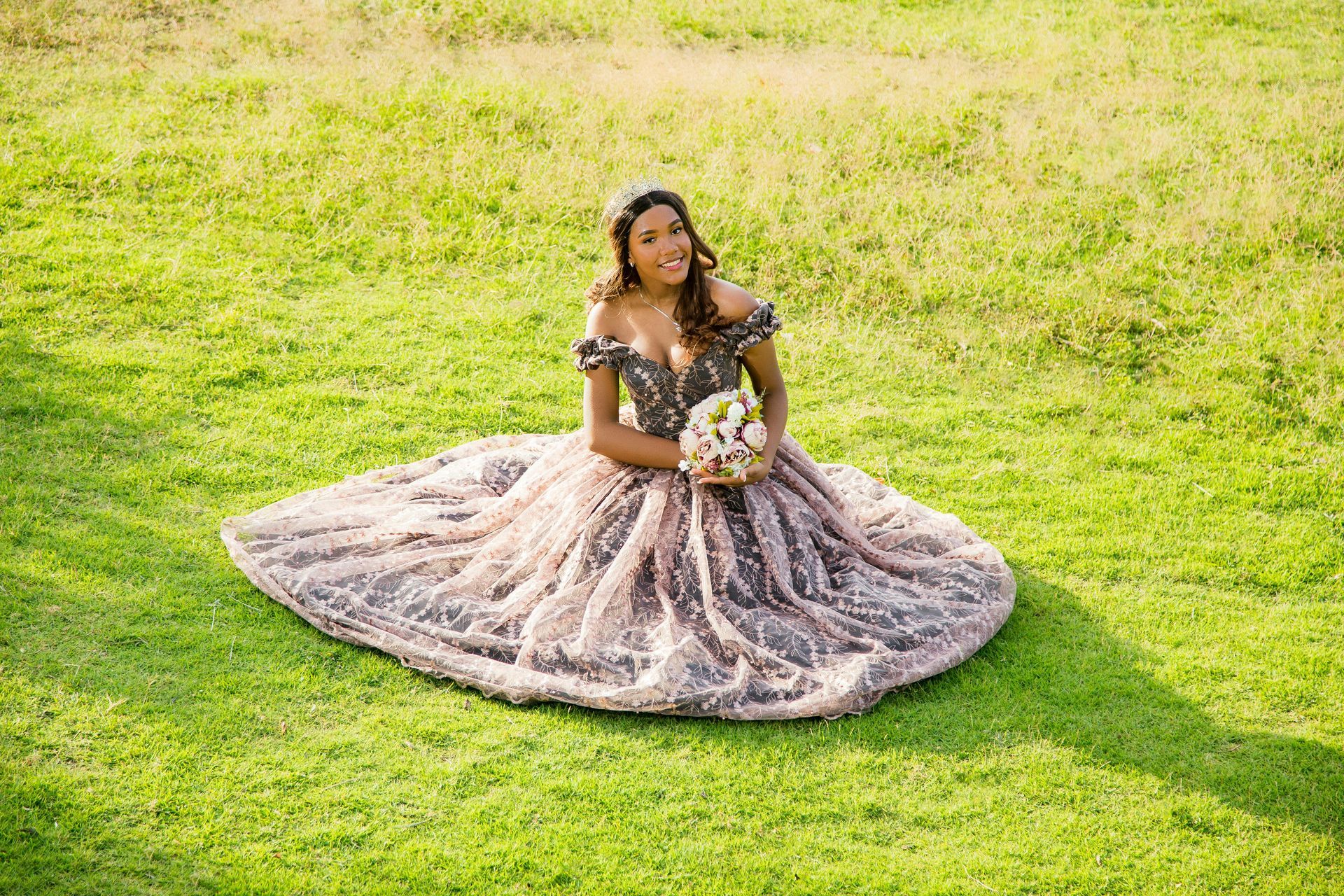 Person in a lavender gown seated on grass, holding a bouquet in a sunlit field