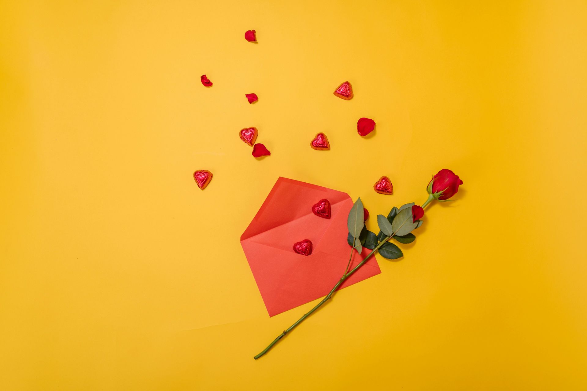 Red rose, chocolate hearts, and red envelope on a yellow background.