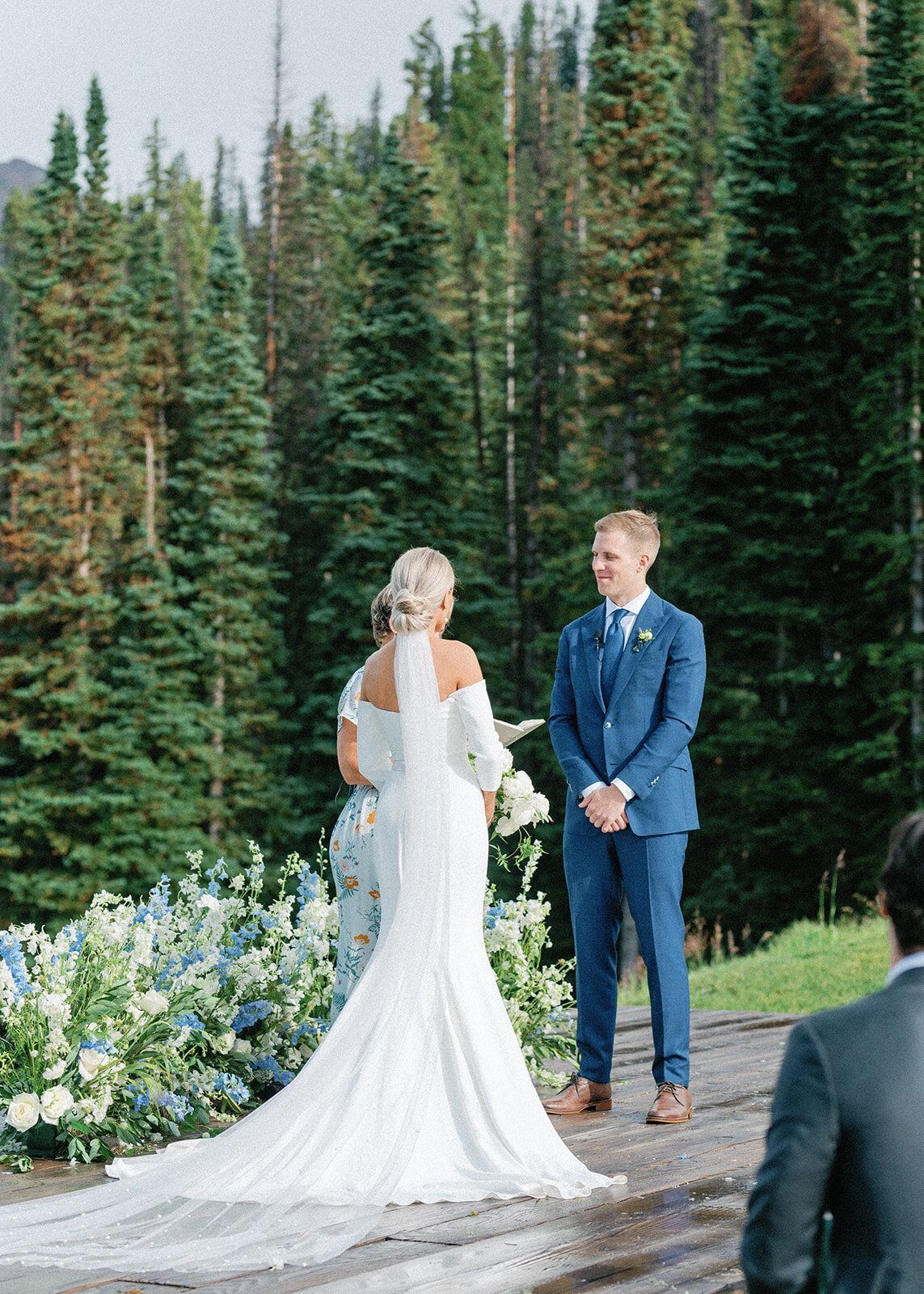 A bride and groom are standing next to each other in front of a forest.