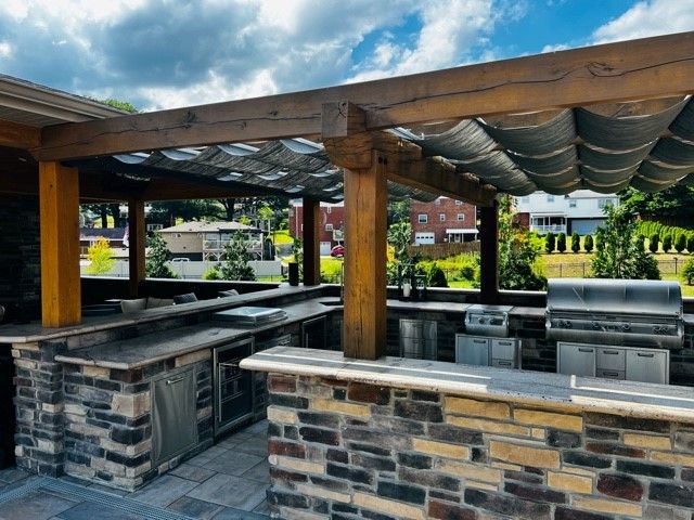 Outdoor kitchen with stone counters, stainless steel appliances, under a shaded pergola on a sunny day.