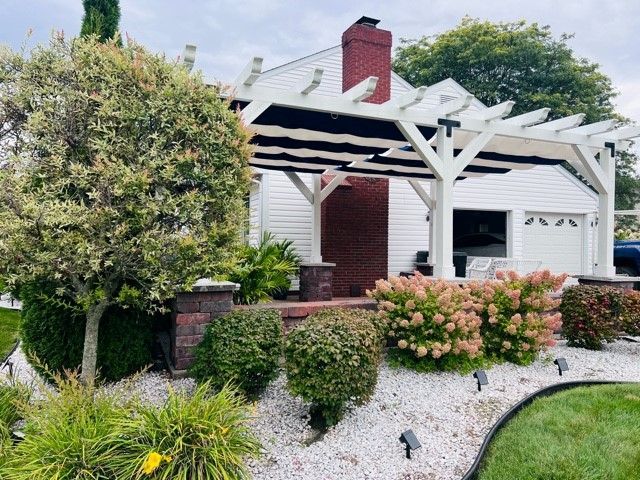 White house with pergola, red brick chimney, and landscaped garden with white rocks and colorful bushes.