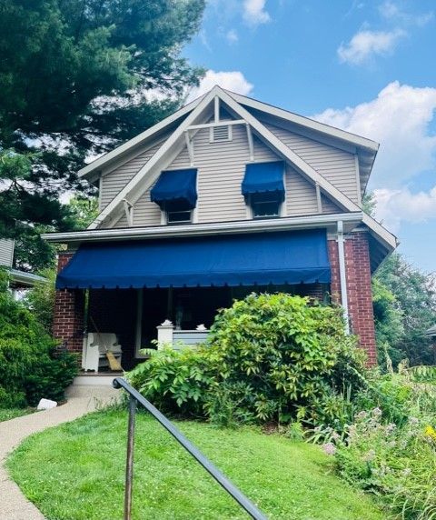 Two-story house with red brick and tan siding, blue awnings over windows and porch, surrounded by greenery.