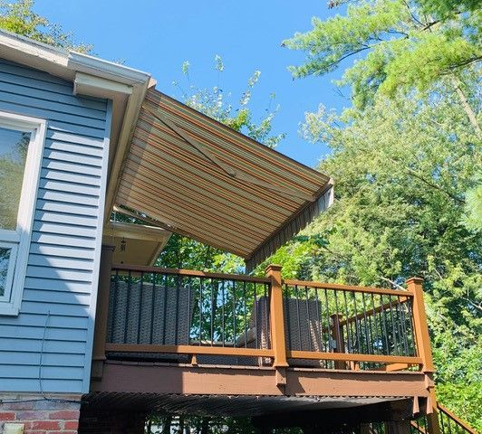 Wooden deck with a beige and brown striped awning extended from a light blue house. Trees in the background.