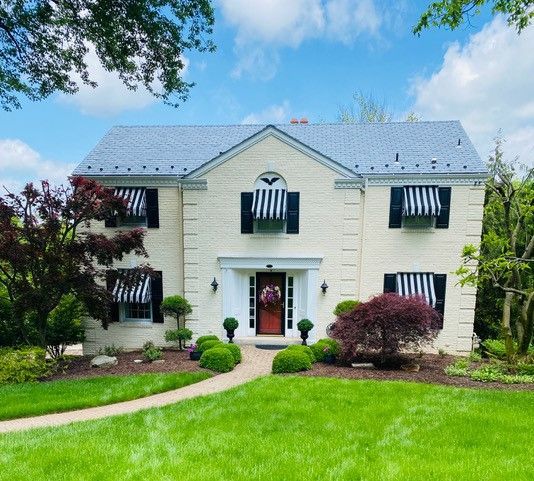 Two-story cream-colored house with black and white striped awnings over windows. Red door, green lawn, blue sky.