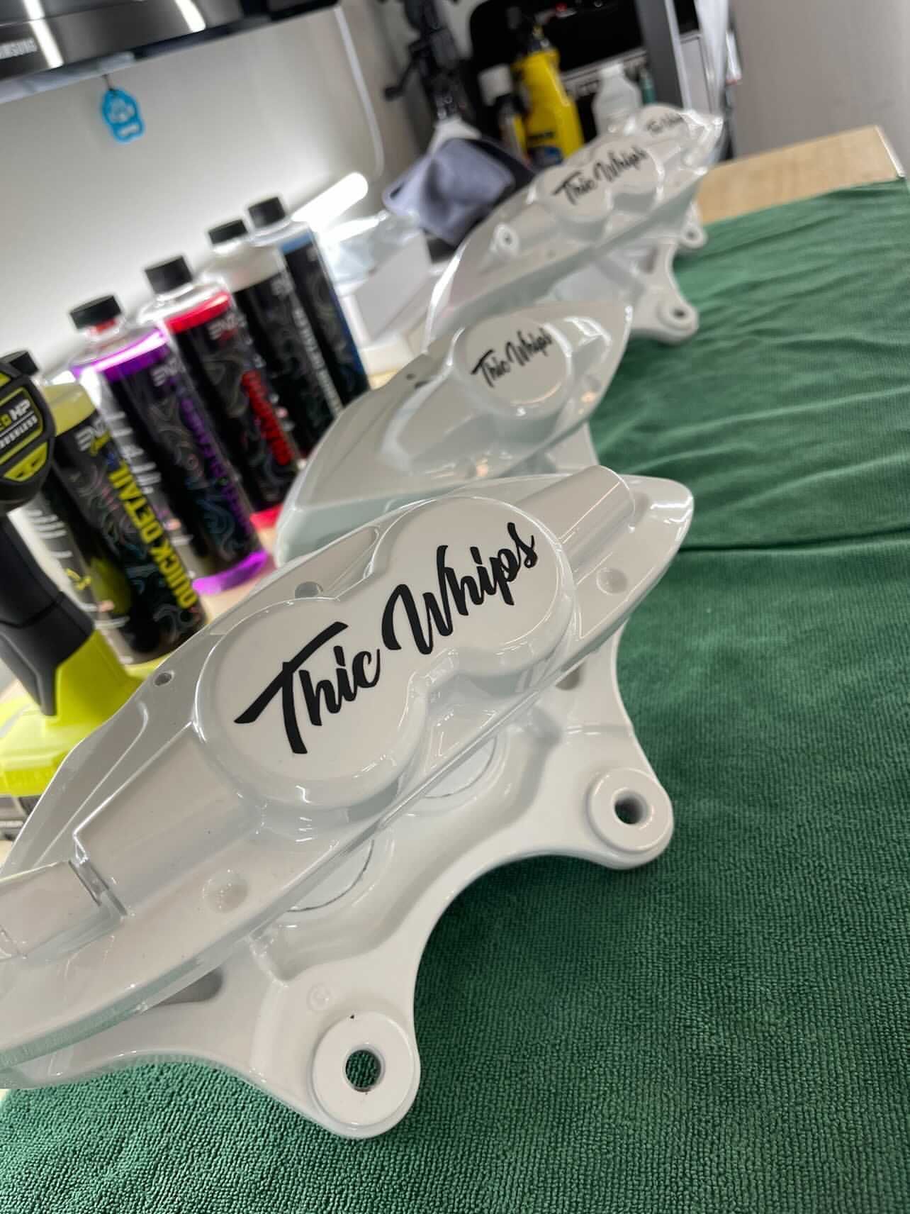 Custom white brake calipers with black script lettering on a workbench with spray cans in the background
