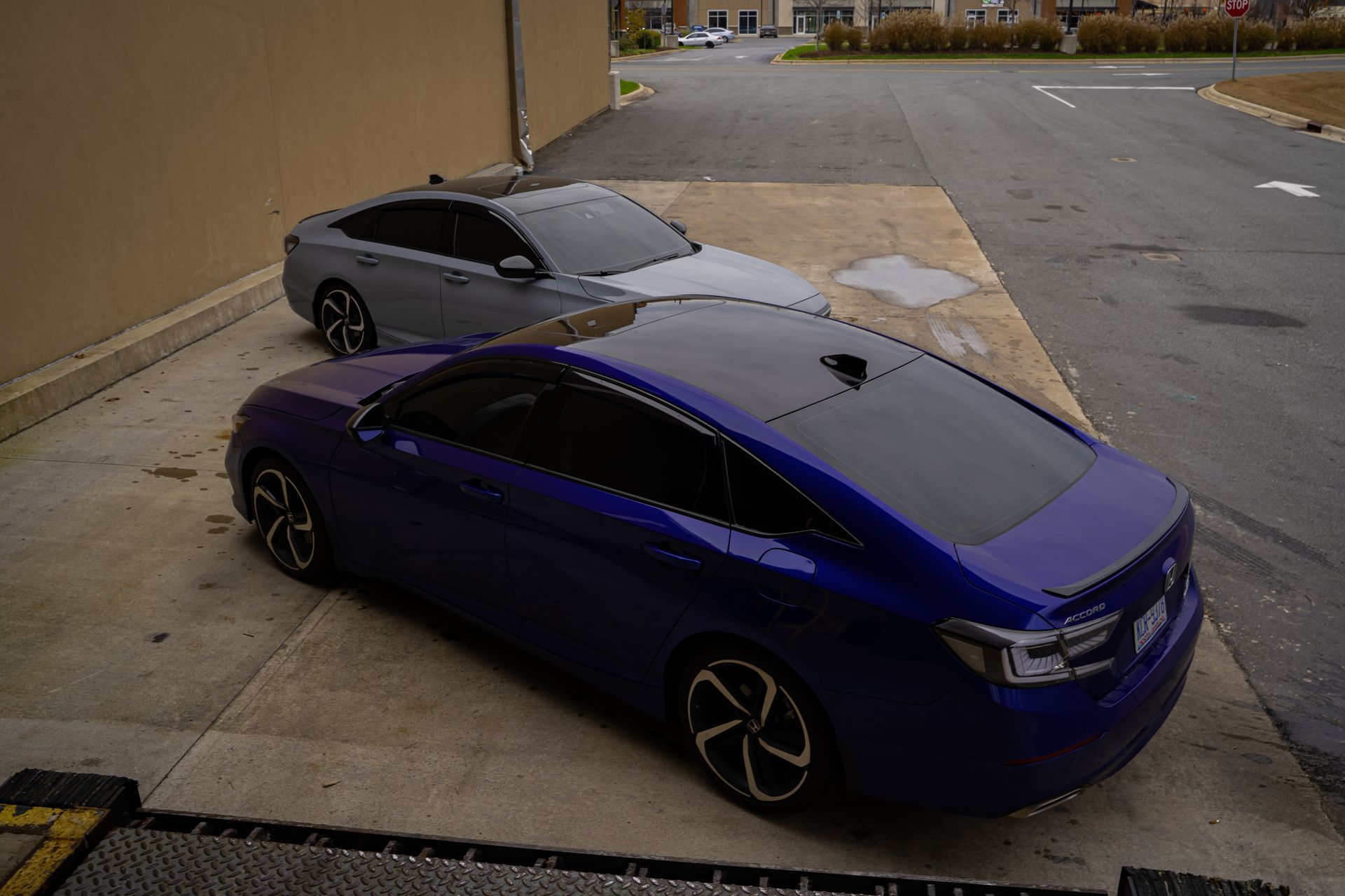 Two parked cars, a blue sedan in front of a gray sedan, beside a building and sidewalk.