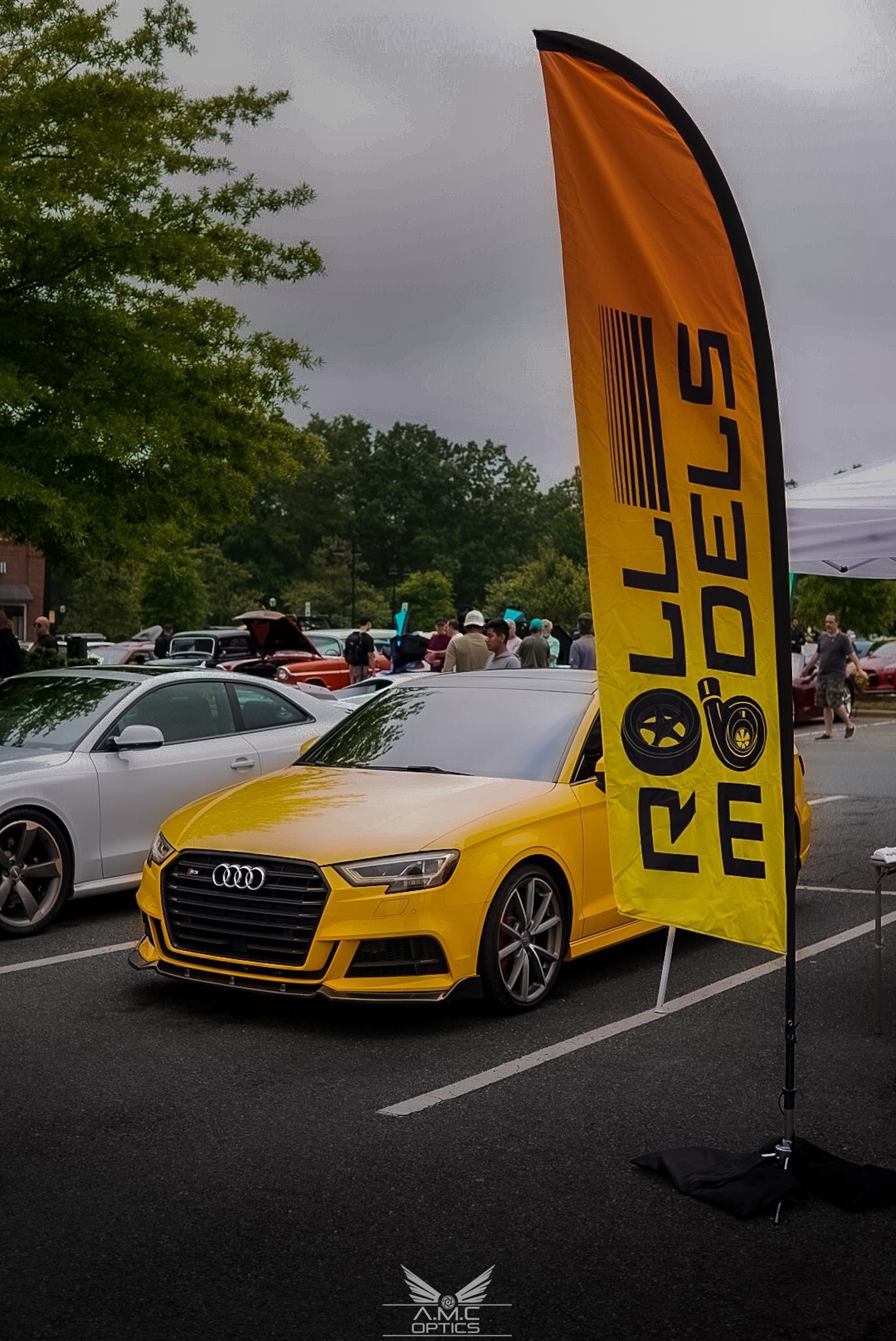 Yellow Audi parked at a rainy outdoor car show beside an orange Jägermeister flag.