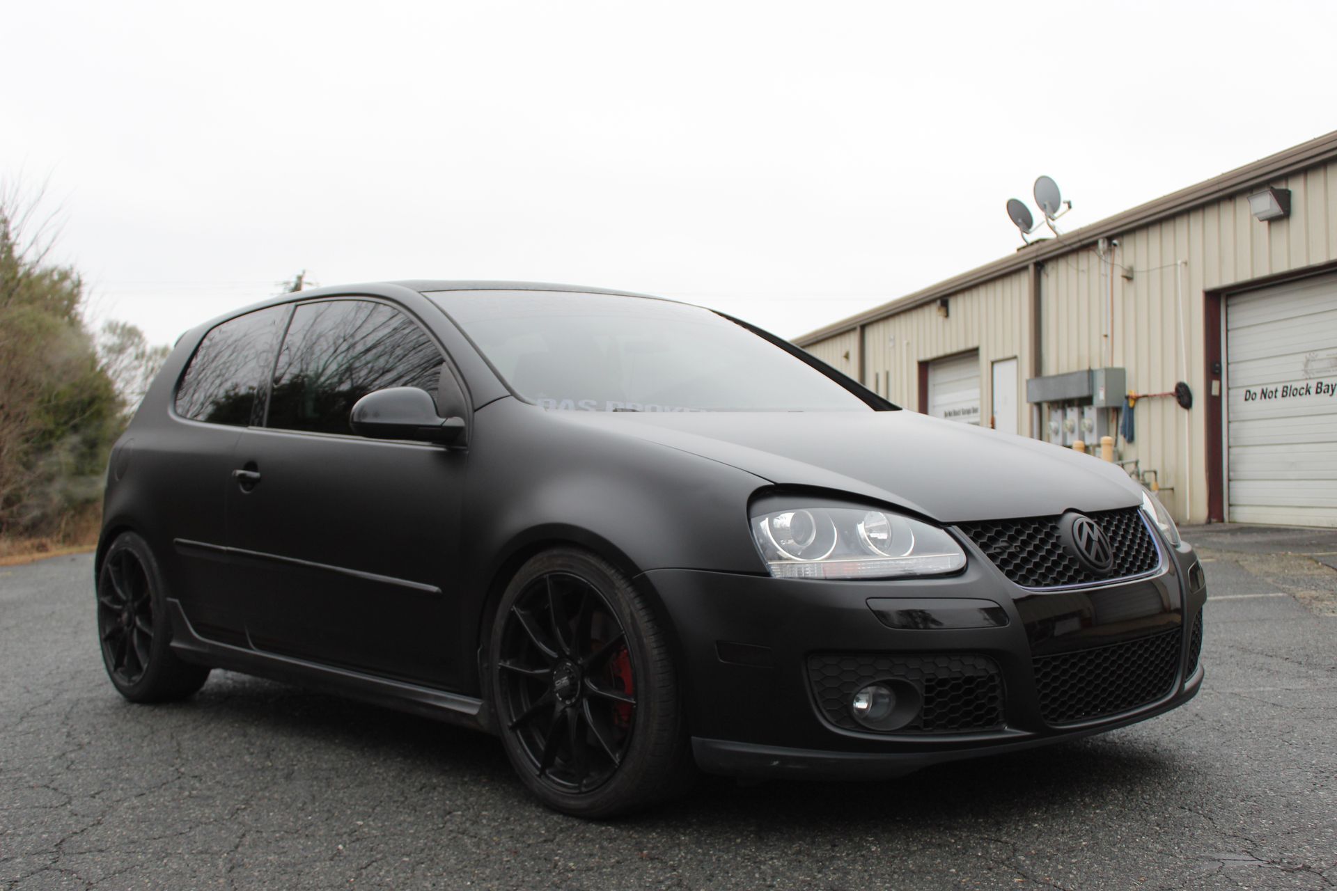 Matte black hatchback parked outside an industrial building on a cloudy day