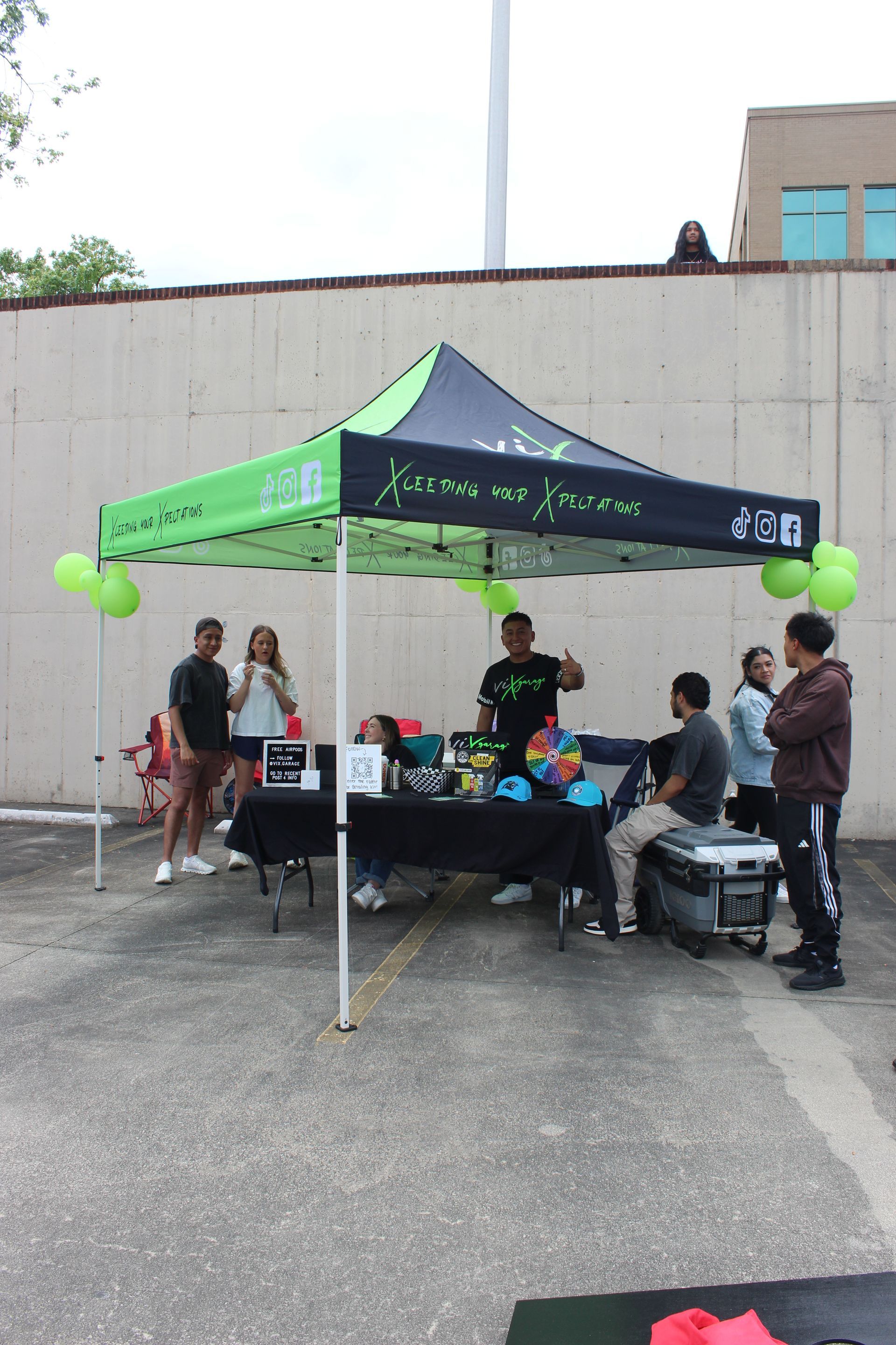 People under a green-and-black canopy tent at an outdoor table with balloons and event materials