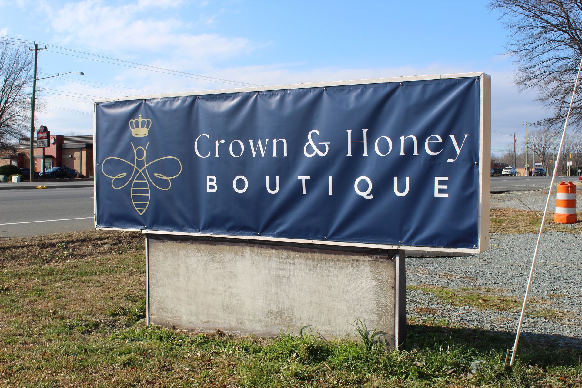 Blue “Crown & Honey Boutique” roadside sign on a concrete base by a grassy shoulder